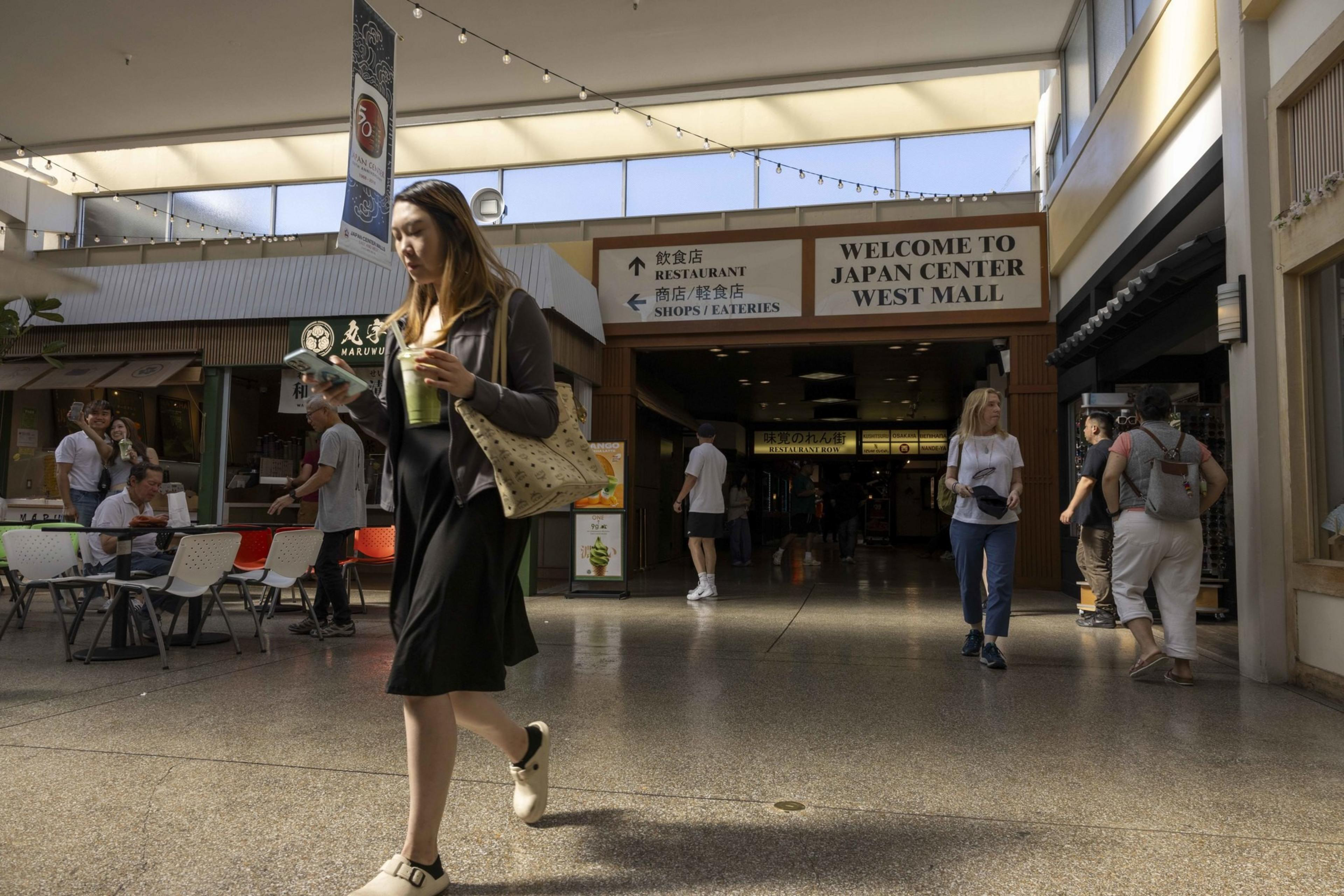 A woman walks through a mall, holding a drink and checking her phone. The sign above reads "Welcome to Japan Center West Mall," with people and shops in the background.