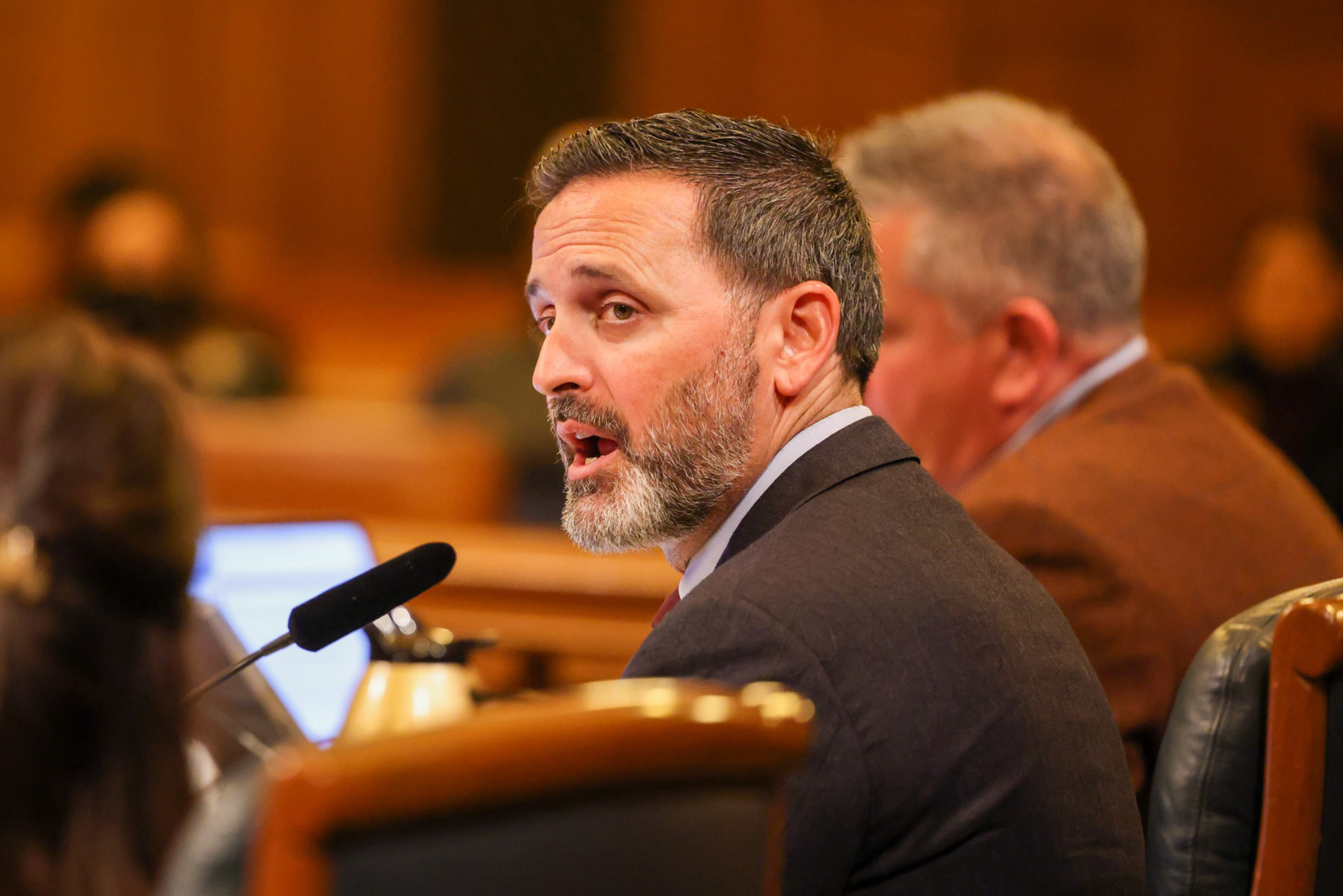 A man with a beard speaks into a microphone in a formal setting with people around.