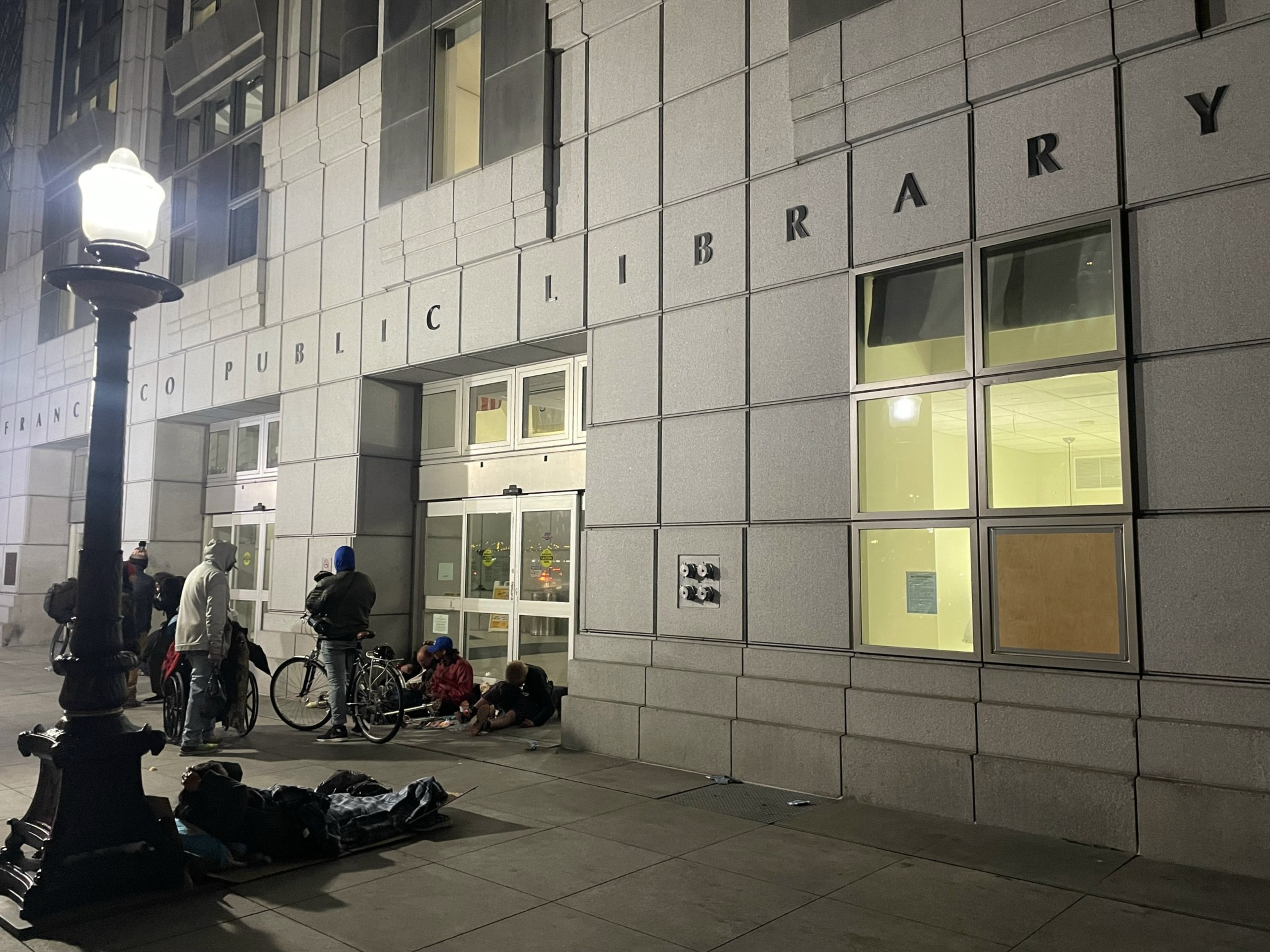 Outside a public library at night, with people gathered by the entrance and others lying on the pavement.