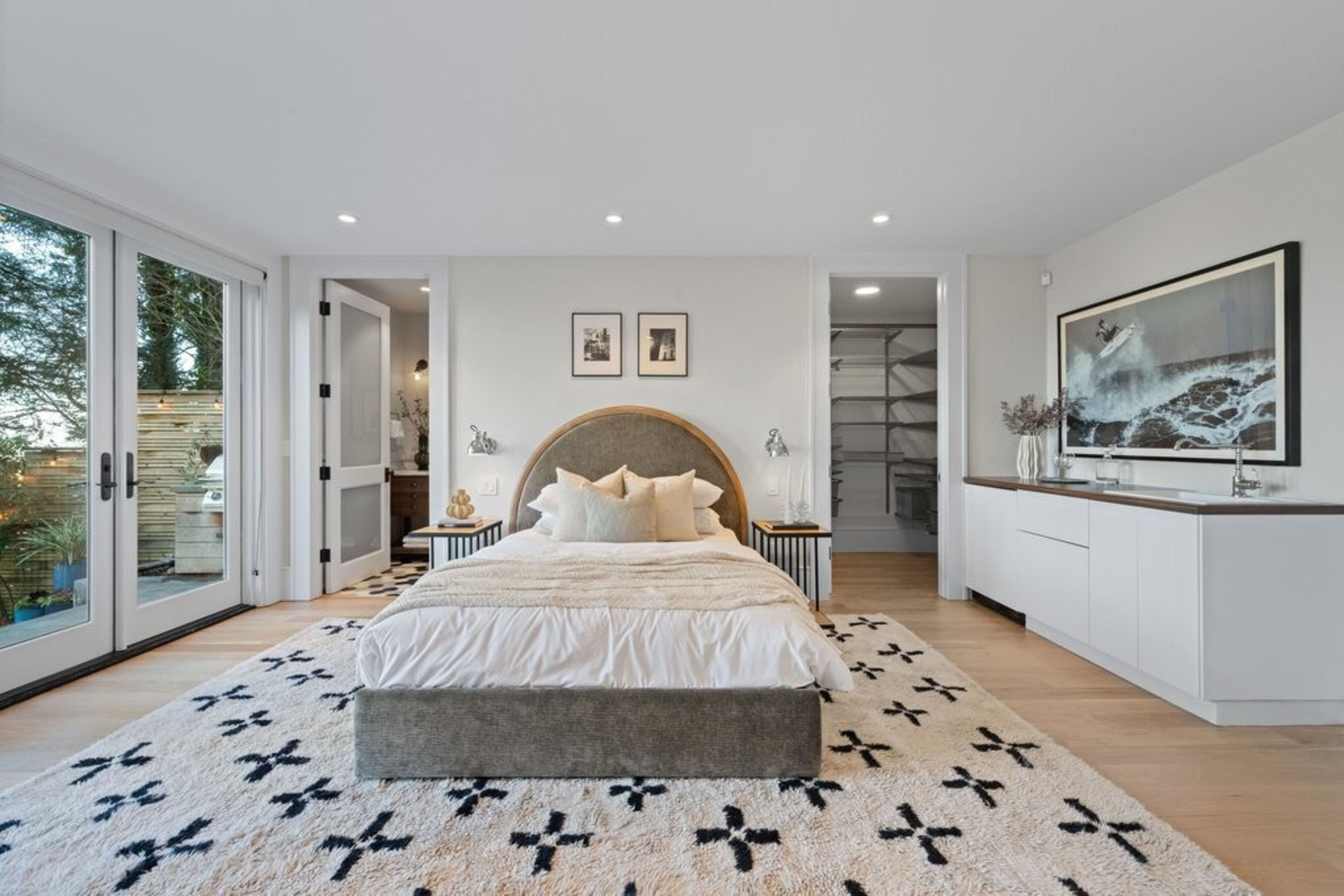 A modern bedroom with a large bed centered on a patterned rug, sliding glass doors to the left, two open doorways, and a white cabinet with a wave painting on the right.