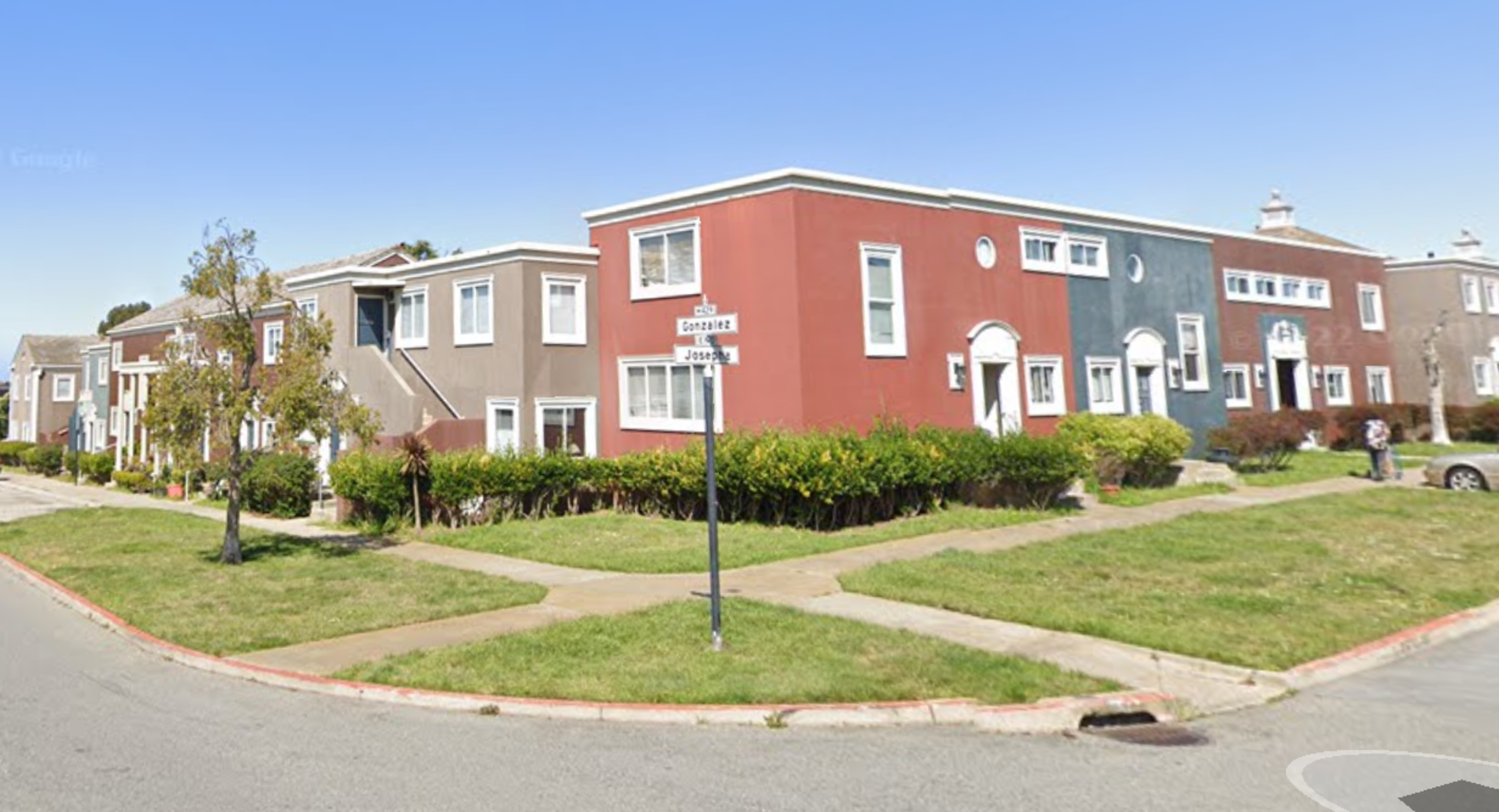Row of colorful two-story houses on a sunny street corner with green lawns and a clear sky.