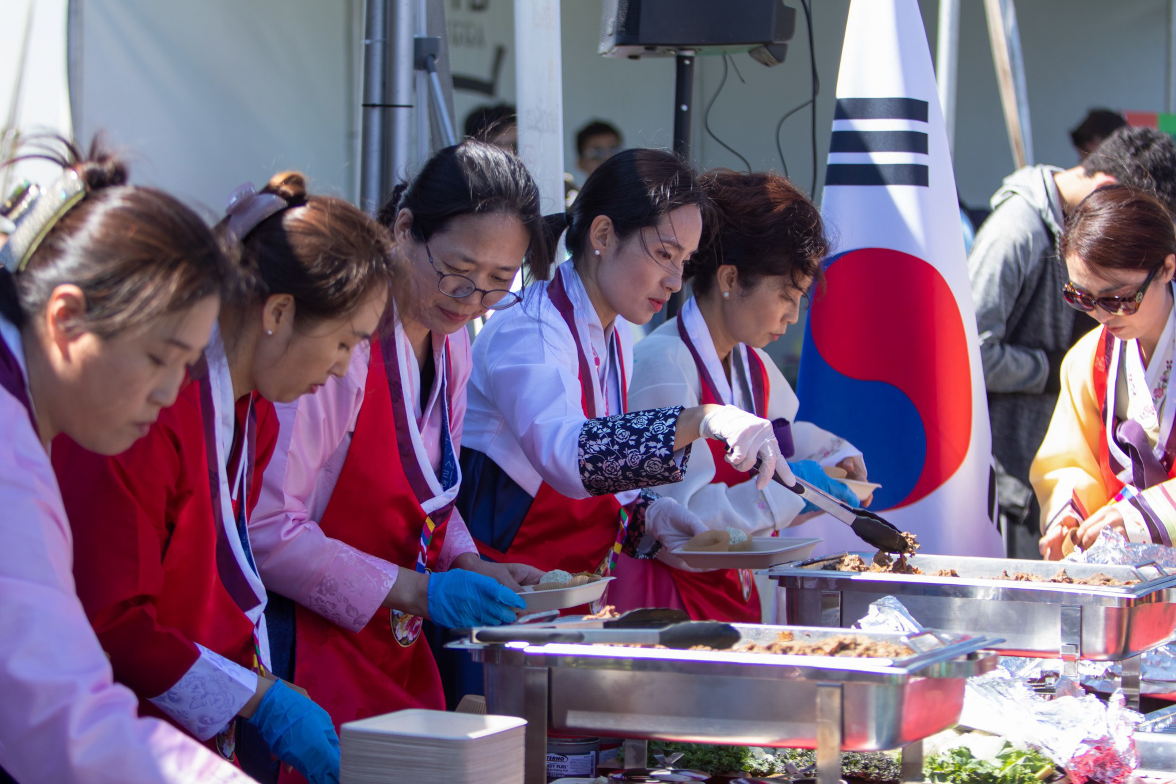 Several women wearing traditional Korean clothing and aprons serve food from large trays at an outdoor event with a South Korean flag in the background.