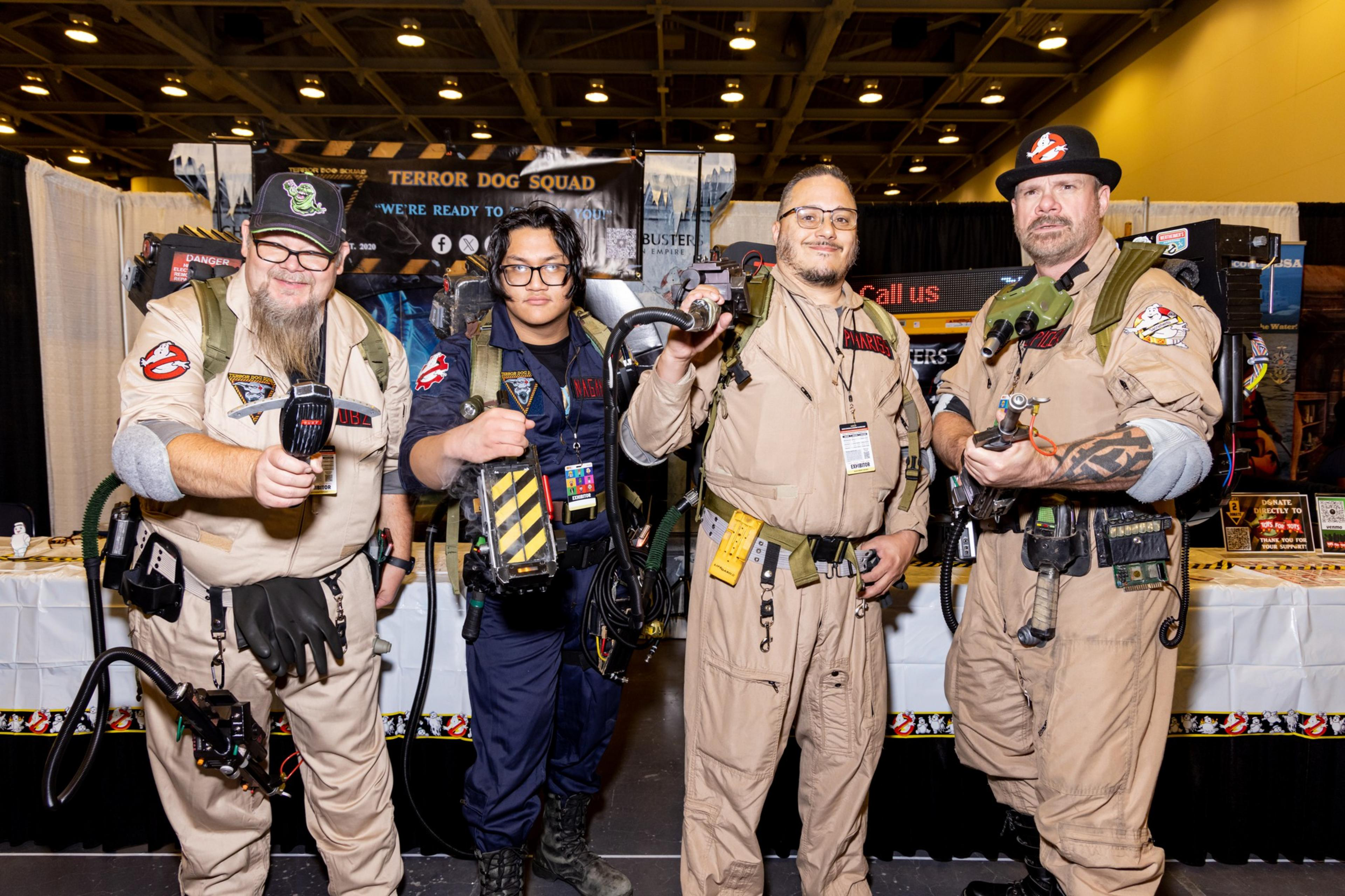 Four people in Ghostbusters costumes stand side by side, holding equipment, in front of a convention booth with a &quot;Terror Dog Squad&quot; banner.