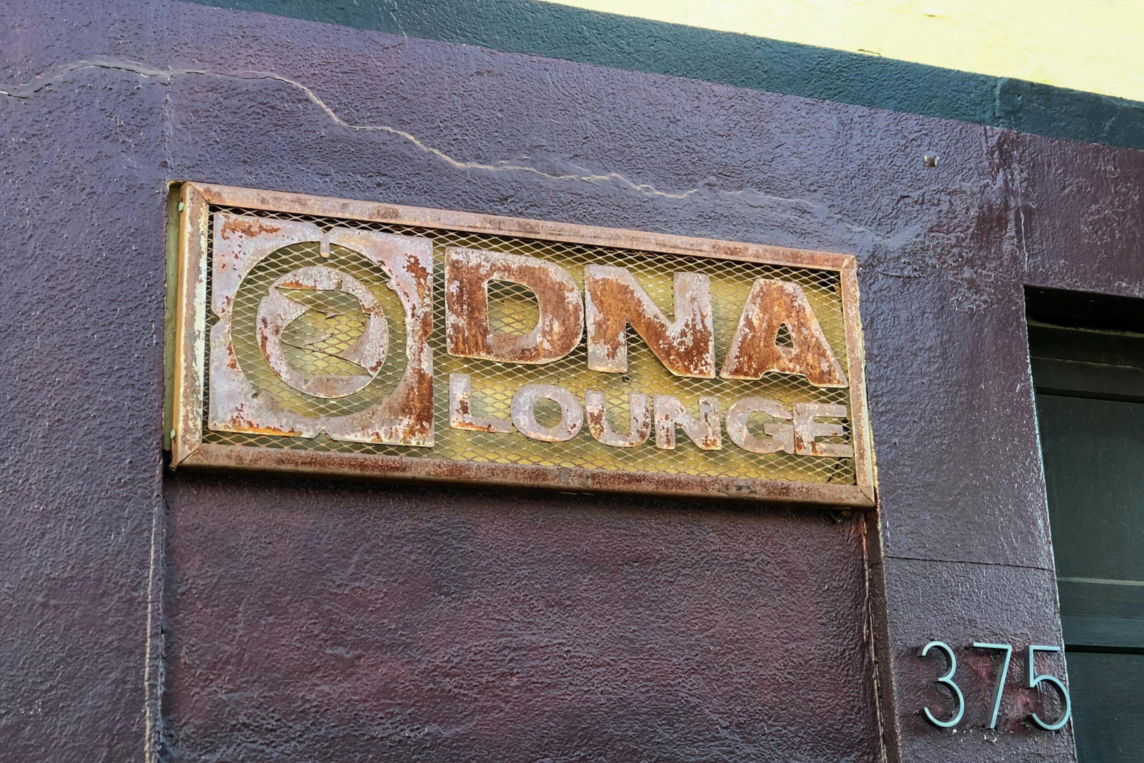A weathered, rusty metal sign reads "DNA LOUNGE" with a circular symbol on a dark purple wall next to the number 375 in metallic numbers.