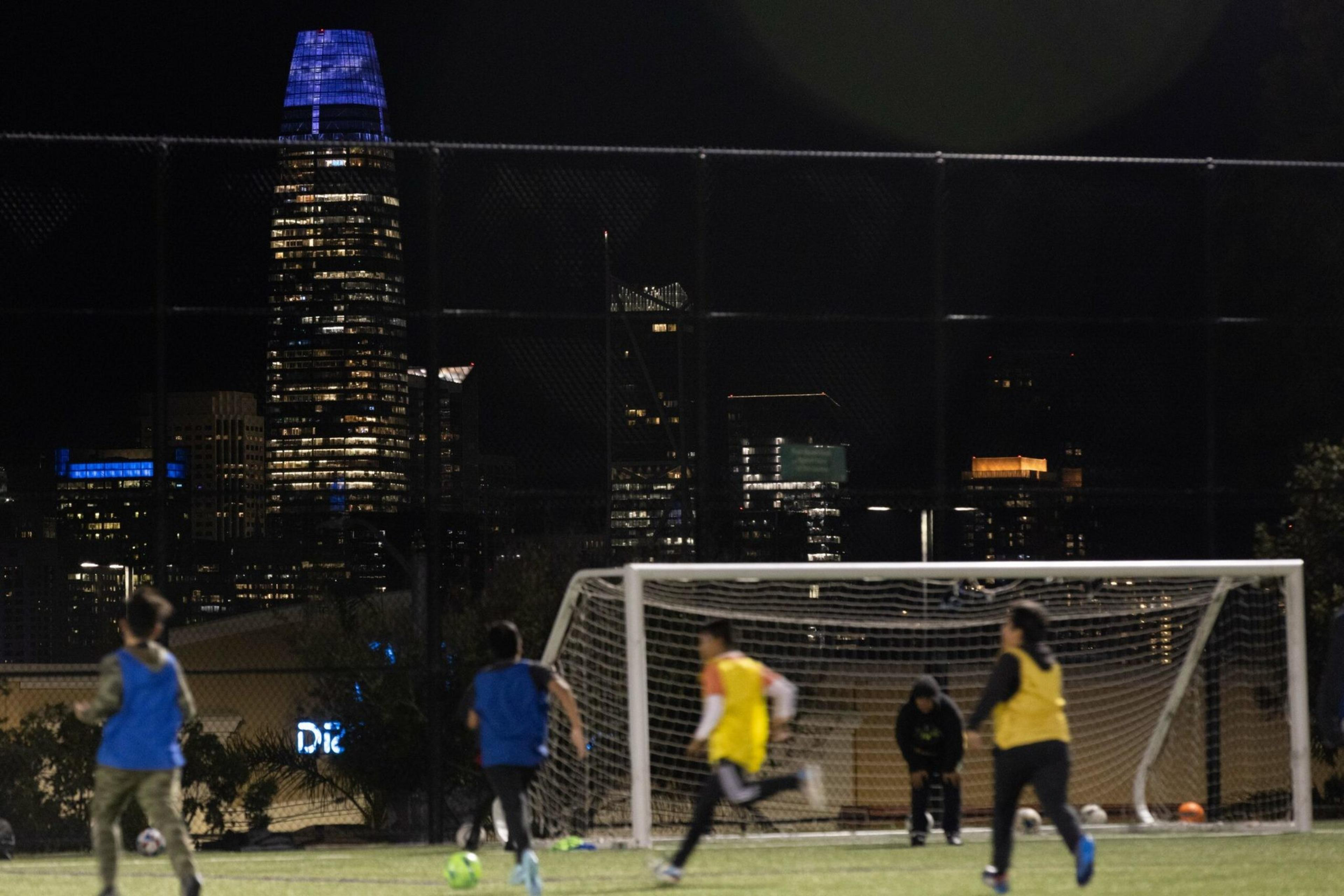 Children play soccer at night on a field, with brightly lit skyscrapers, including a building illuminated in blue, visible in the city skyline behind them.
