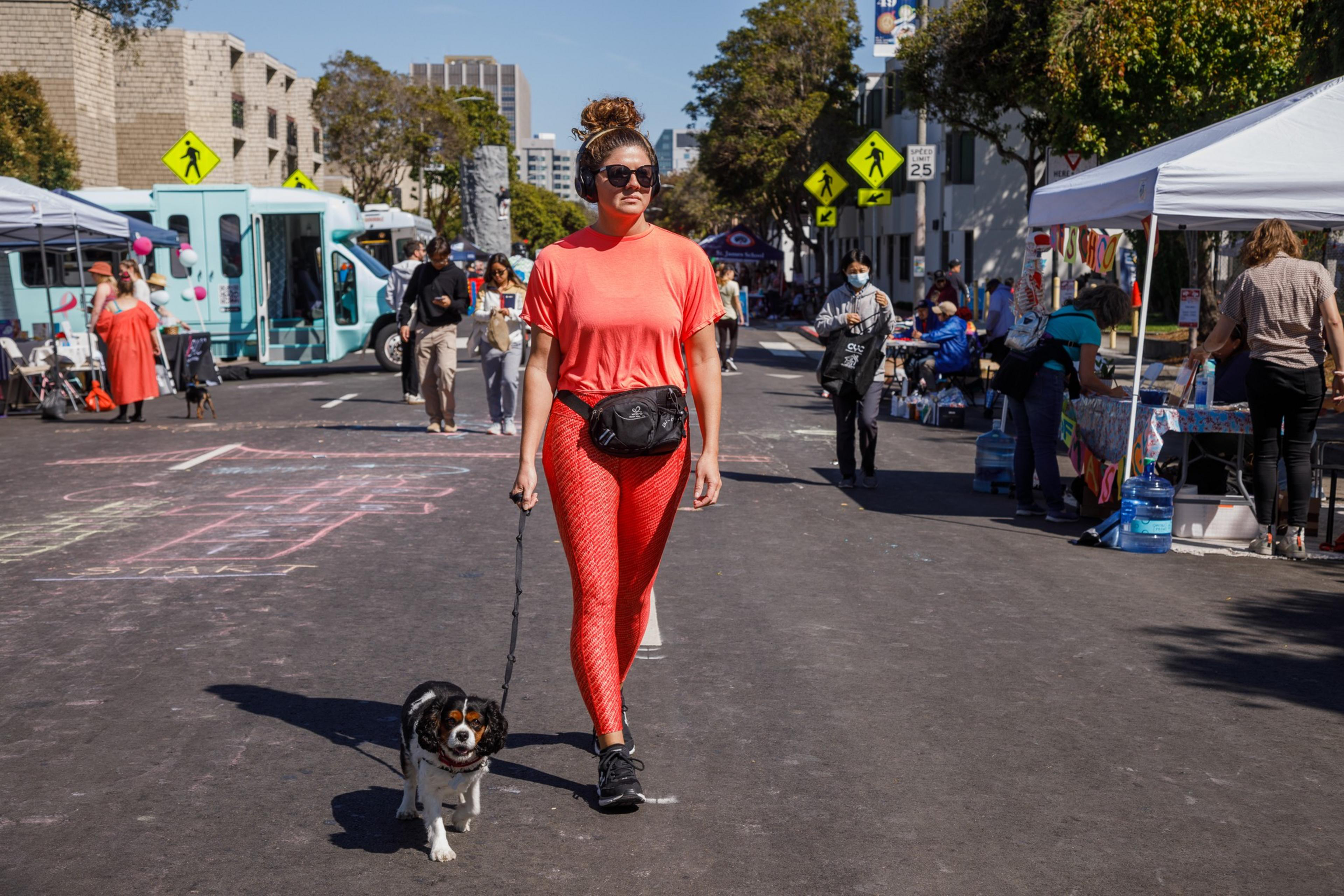 A woman in a bright orange outfit walks a small dog along a sunny street market, with stalls and people around.
