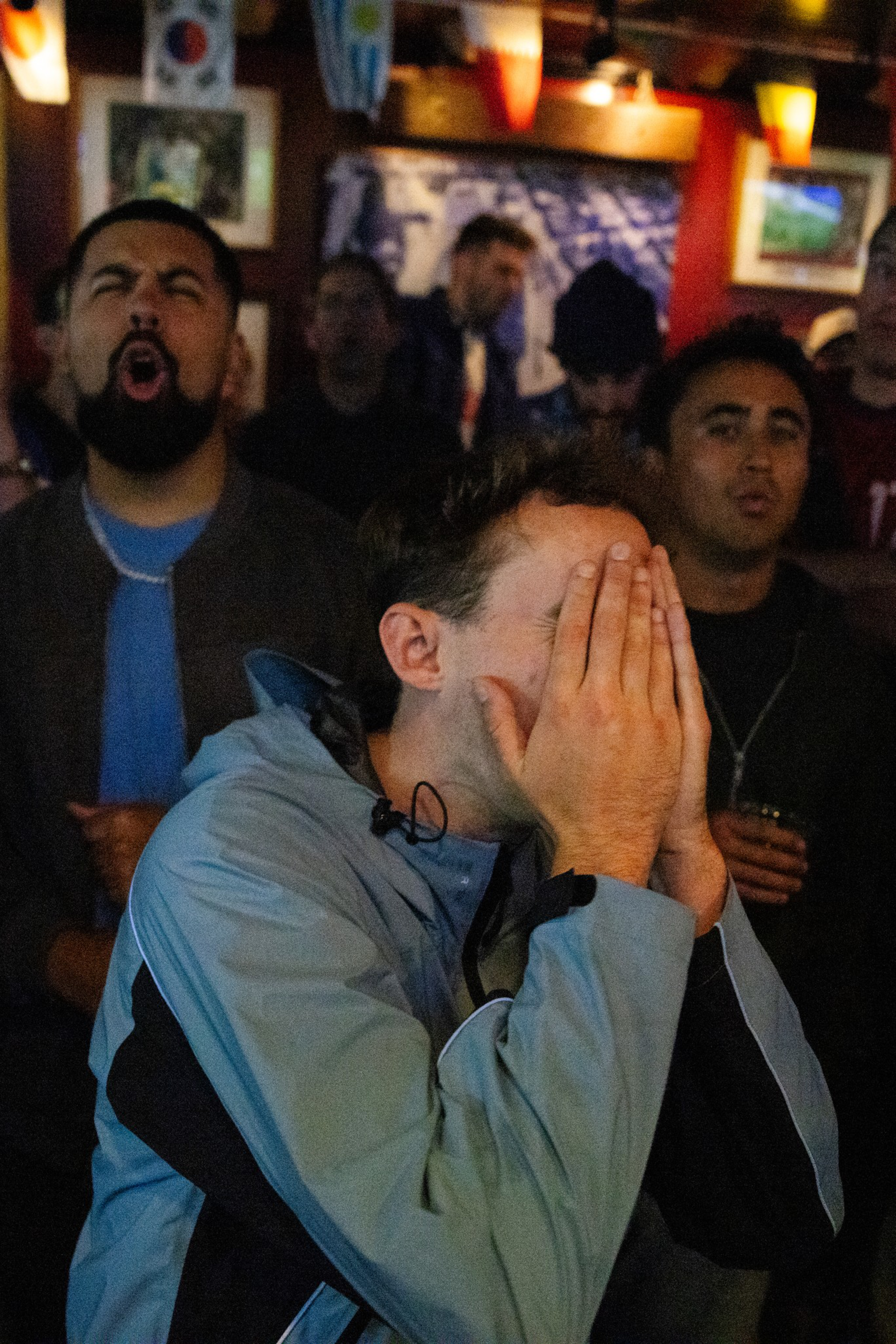 A man in a light blue jacket covers his face with his hands, while others behind him show surprise or frustration, likely watching a sports event.