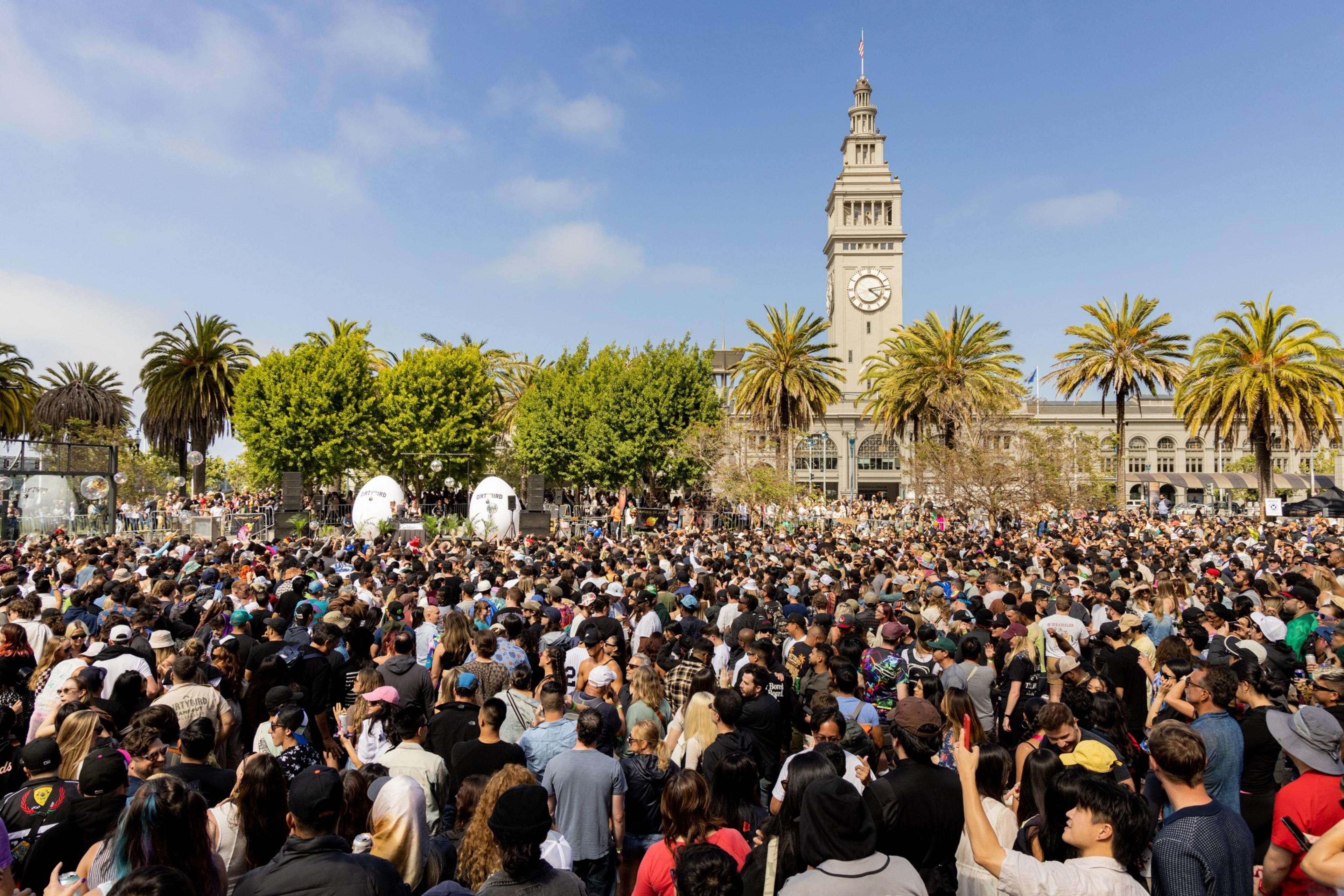 A large crowd gathers outdoors under a sunny sky with palm trees nearby. In the background, a clock tower overlooks the scene, indicating a public event.