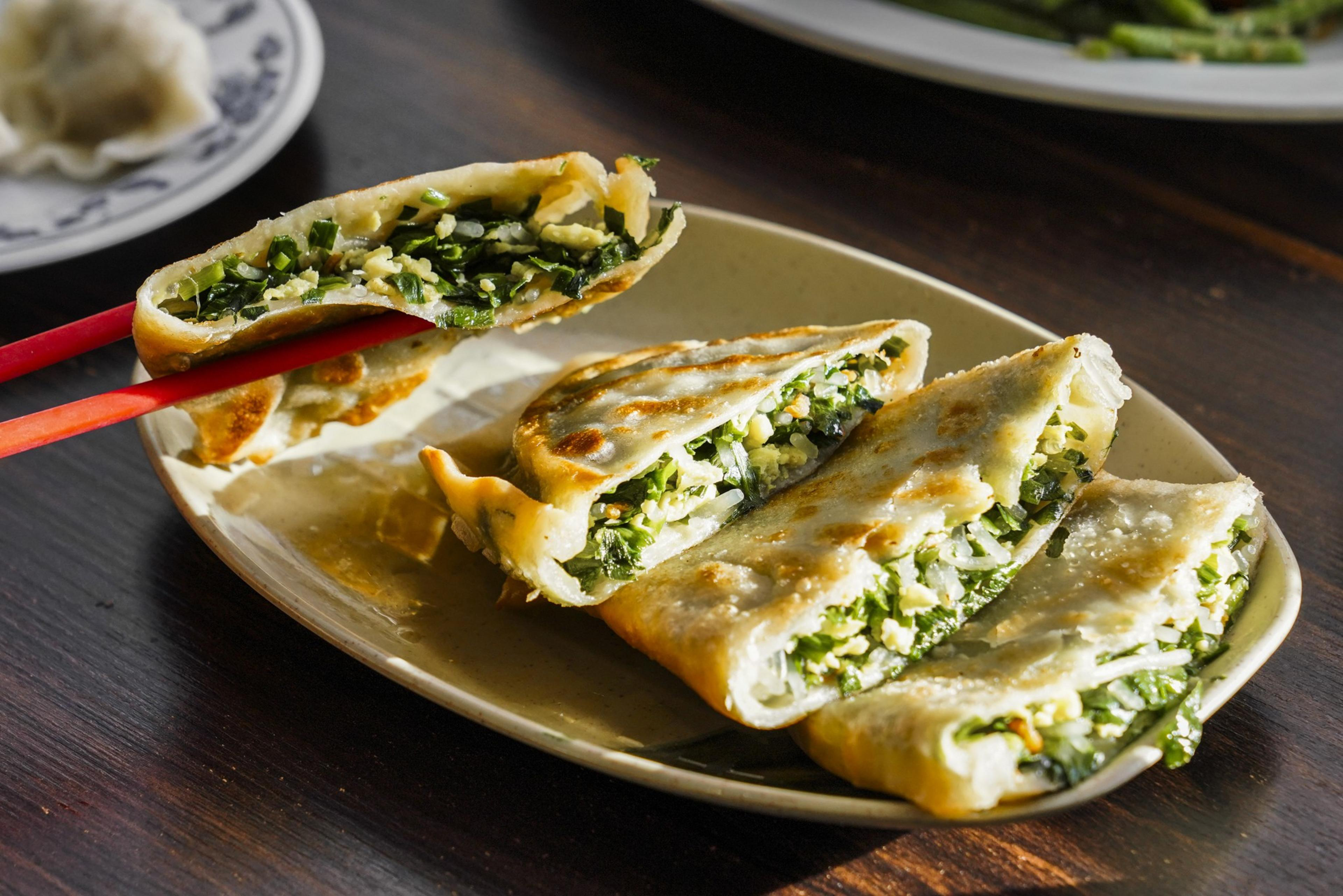 A plate of pan-fried dumplings filled with green vegetables and perhaps egg, with one piece held by red chopsticks over a wooden table.