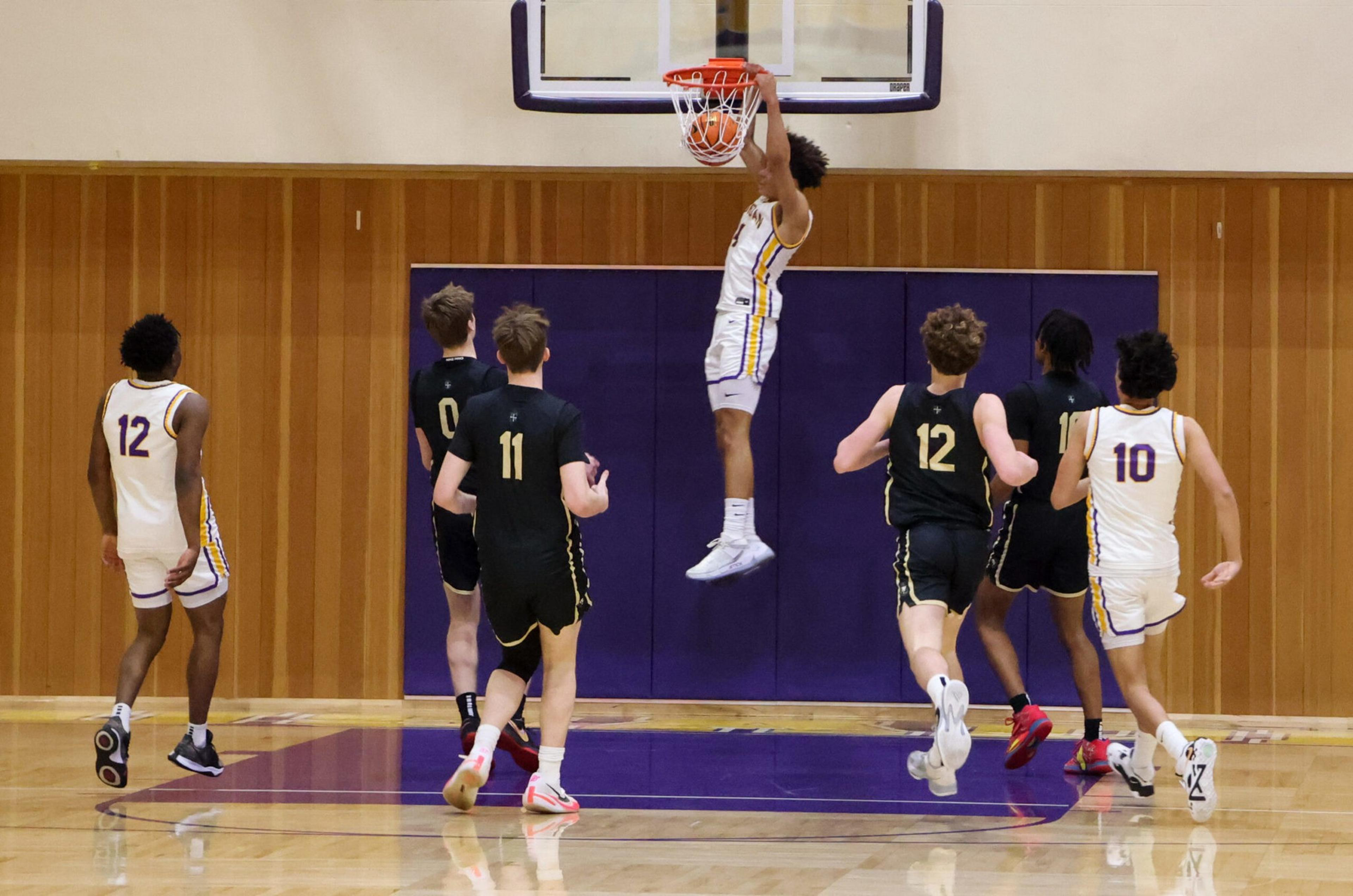 A basketball player in a white uniform jumps to dunk the ball while six other players, three in white and three in black uniforms, watch or run nearby.