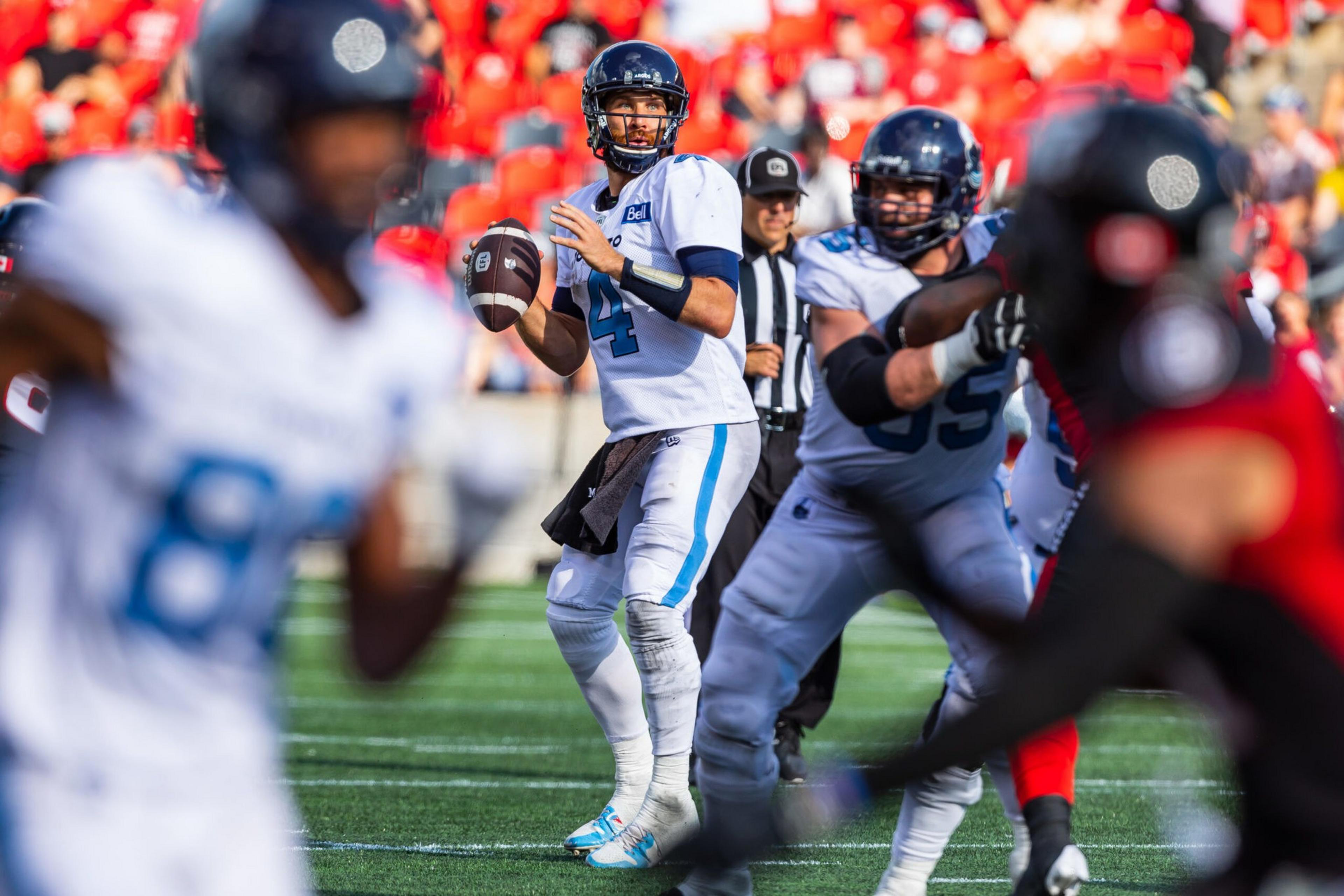 A football quarterback, wearing a blue-and-white uniform, prepares to throw a pass while teammates block opposing players in red and black.