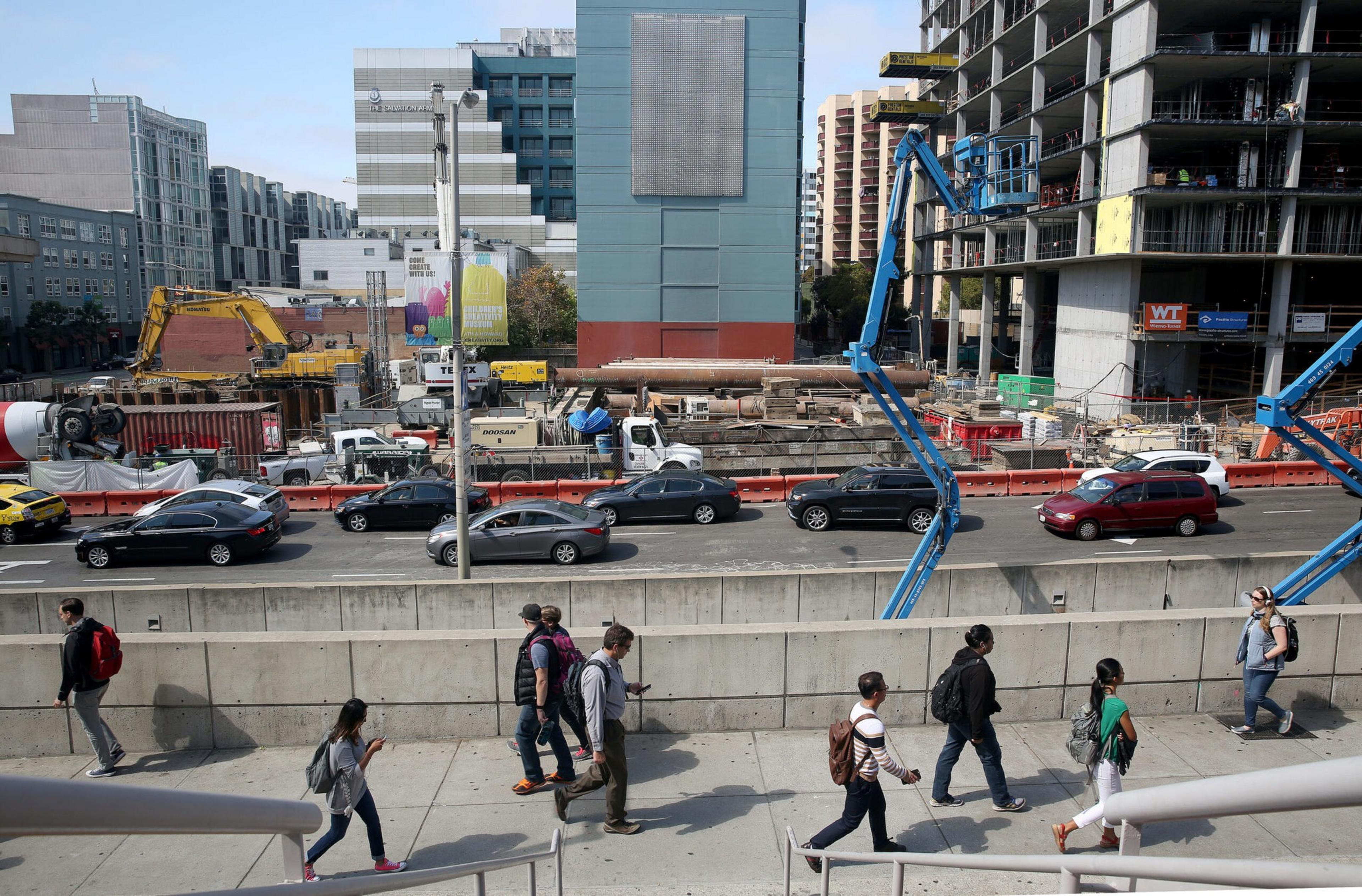 Pedestrians walk along a sidewalk beside a busy road with parked cars and an active construction site in the background, surrounded by tall buildings.