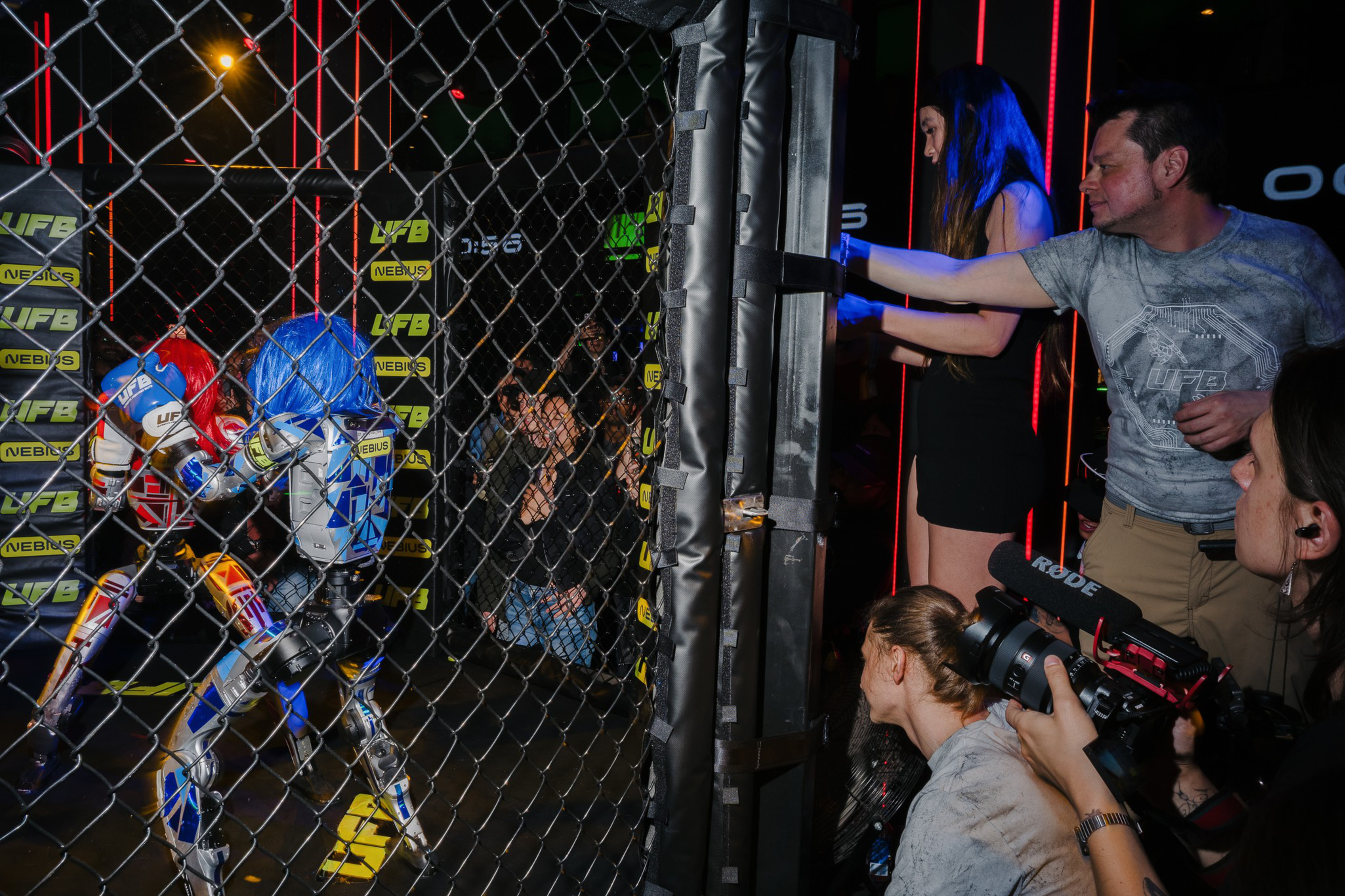 Two robot fighters are engaged inside a cage while several people outside film and cheer them on in a dimly lit venue with neon lights.