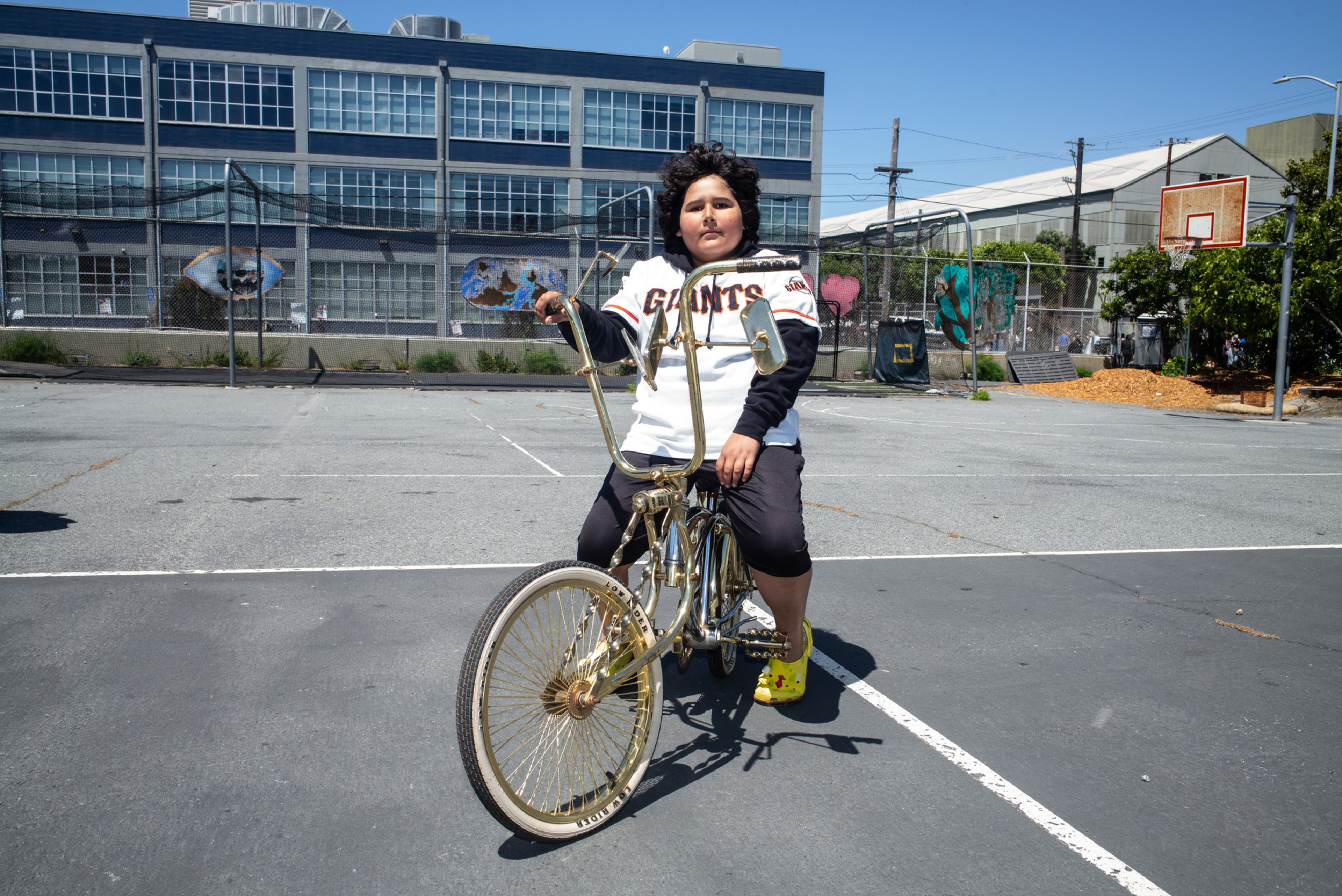 A boy wearing a Giants jersey and yellow Crocs rides a gold lowrider bike on a basketball court in front of a building with large windows.