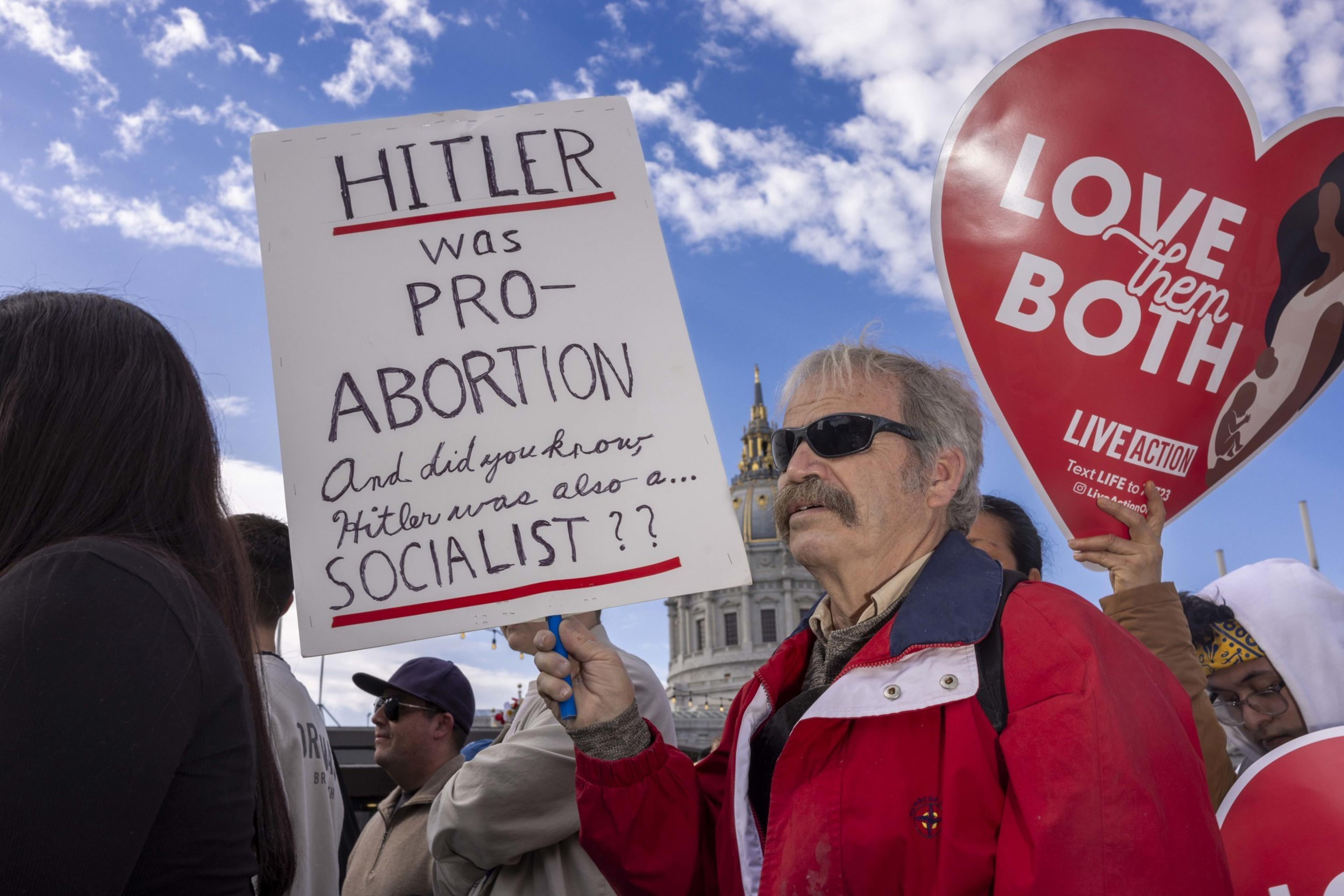 A man in a red jacket holds a sign about Hitler and socialism during a protest. Another person holds a heart-shaped sign saying &quot;Love them both,&quot; against a clear sky.