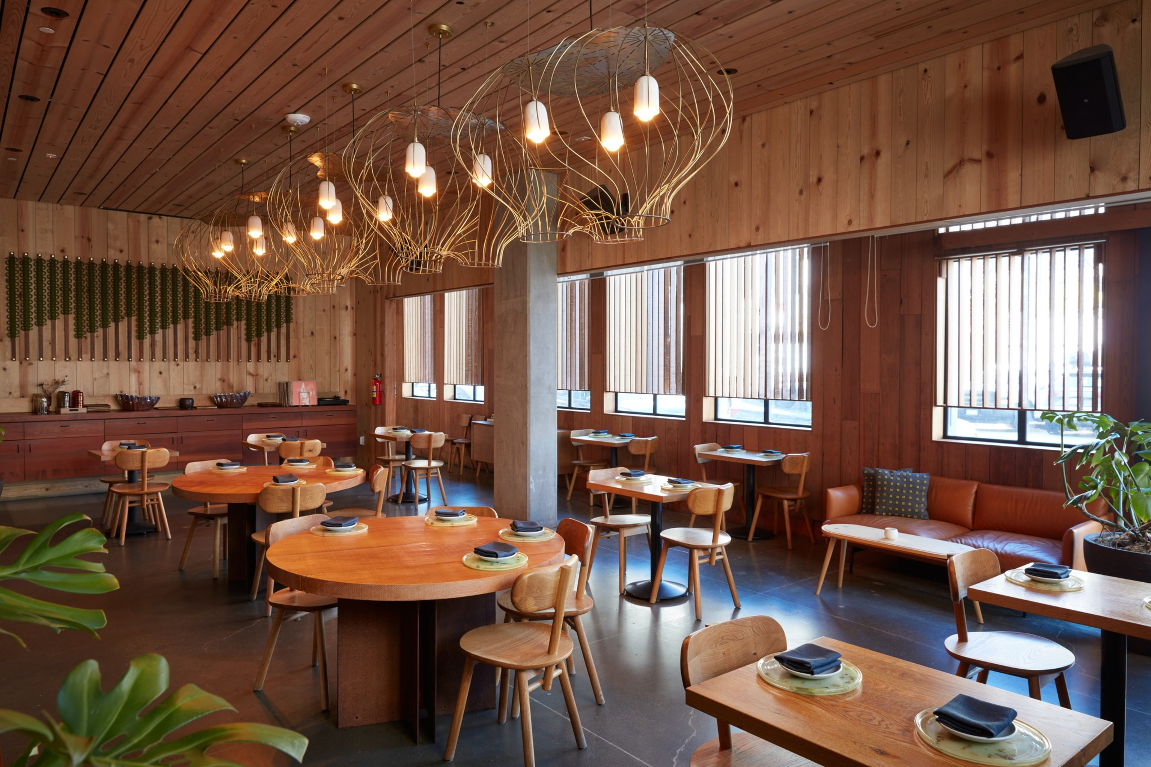 The image shows a stylish restaurant with wooden furniture, round tables, pendant lights, and large windows. A couch is along one wall, adding a cozy touch.