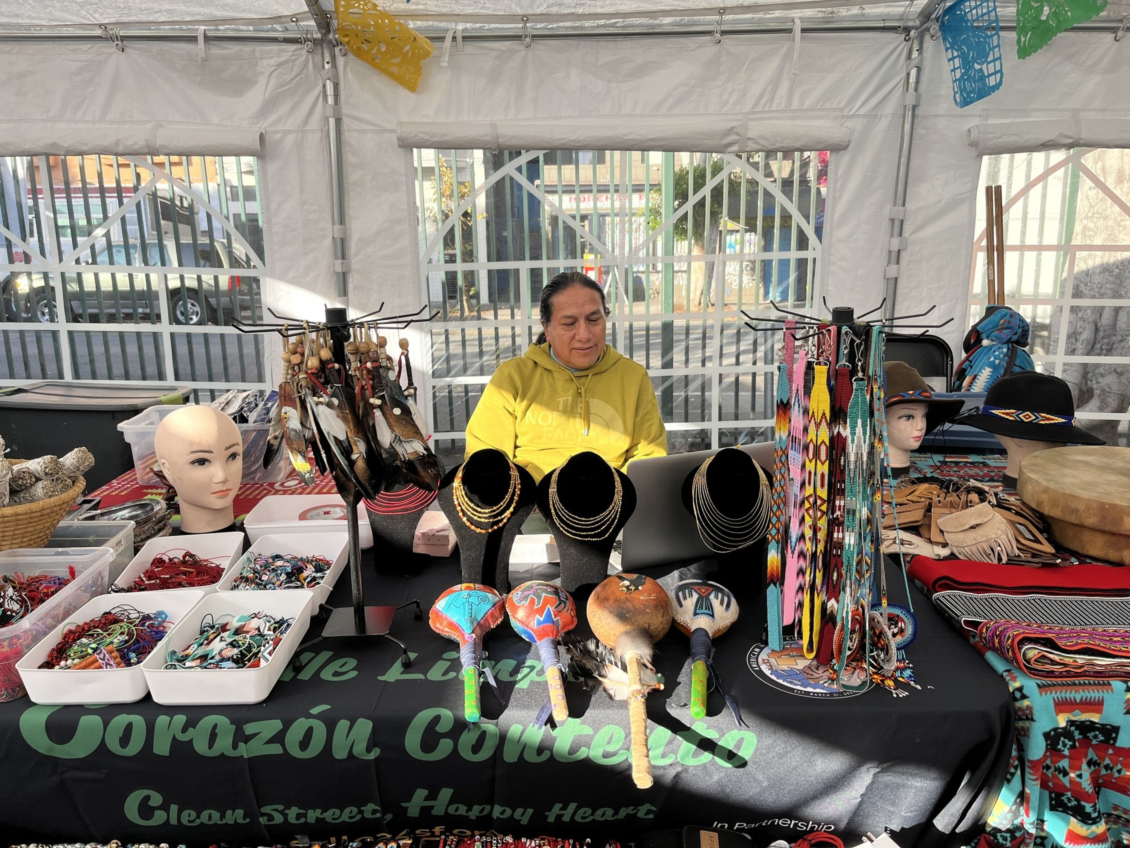 A man in a yello shirt sits behind a table with variosu good including barcelets and necklaces