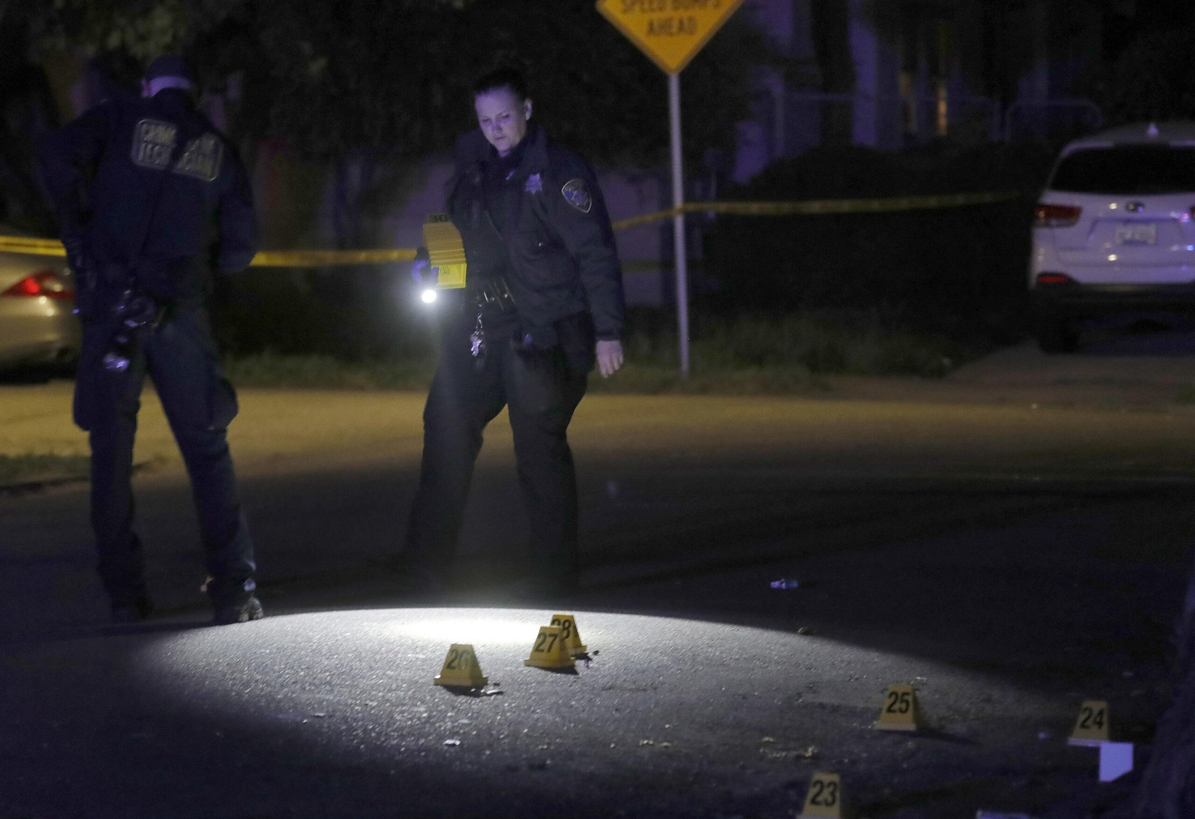 A police officer uses a flashlight to examine numbered evidence markers on a dimly lit street at night.