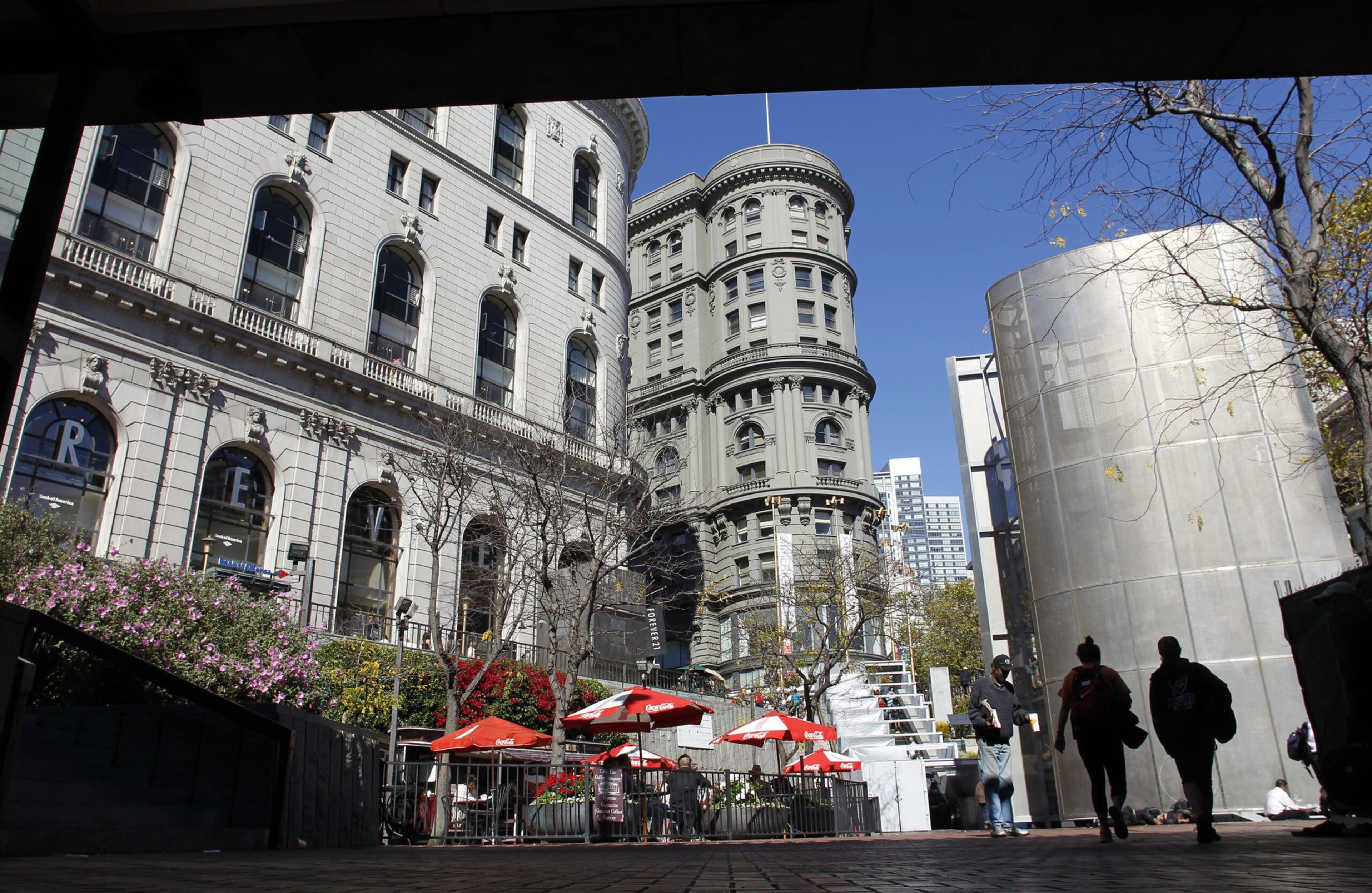 A city plaza with curved historic buildings, red umbrellas shading outdoor seating, leafless trees, and people walking near a modern cylindrical structure.