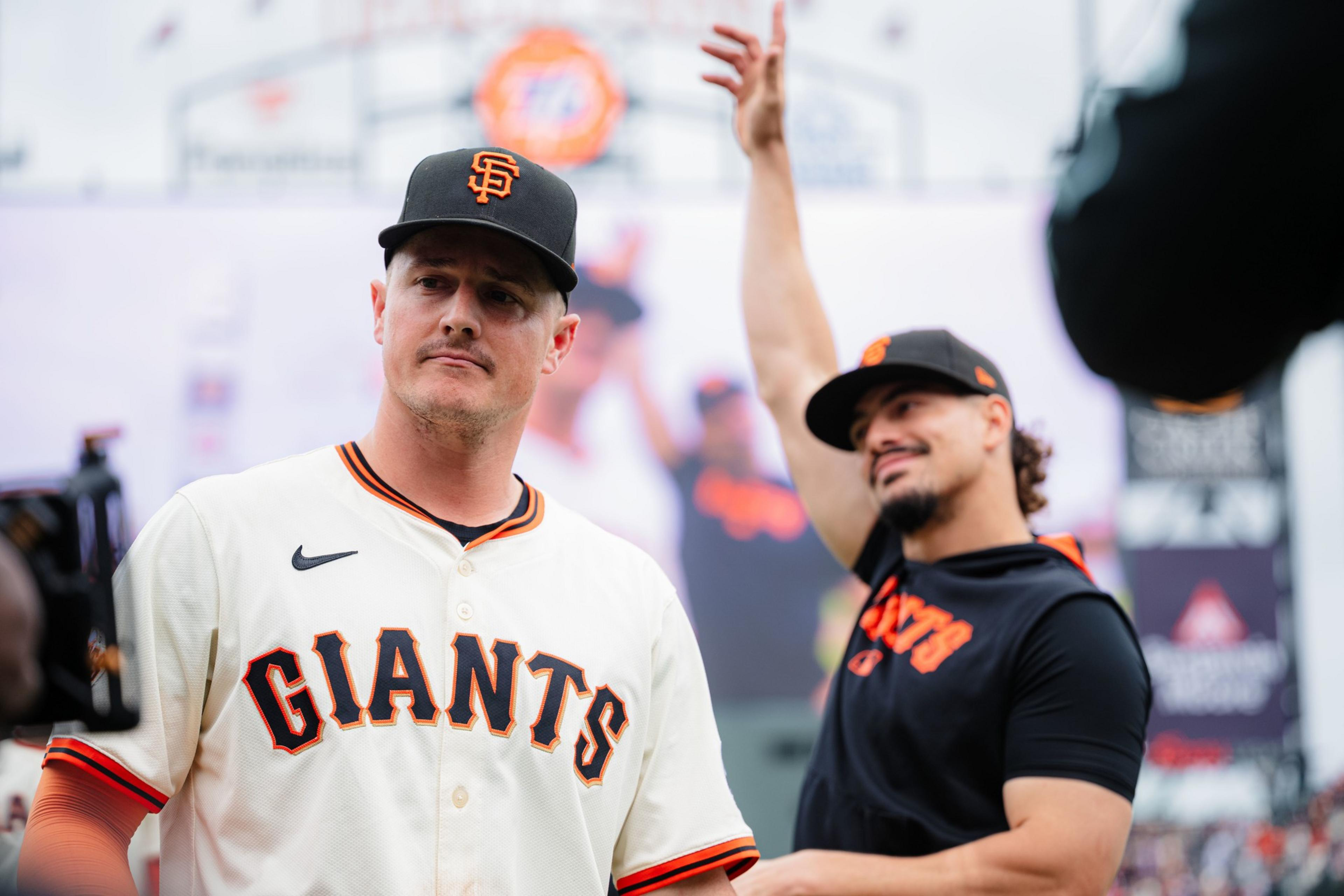 A man in a white San Francisco Giants baseball jersey and cap looks forward seriously while another man in a black Giants shirt and cap smiles and waves behind him.
