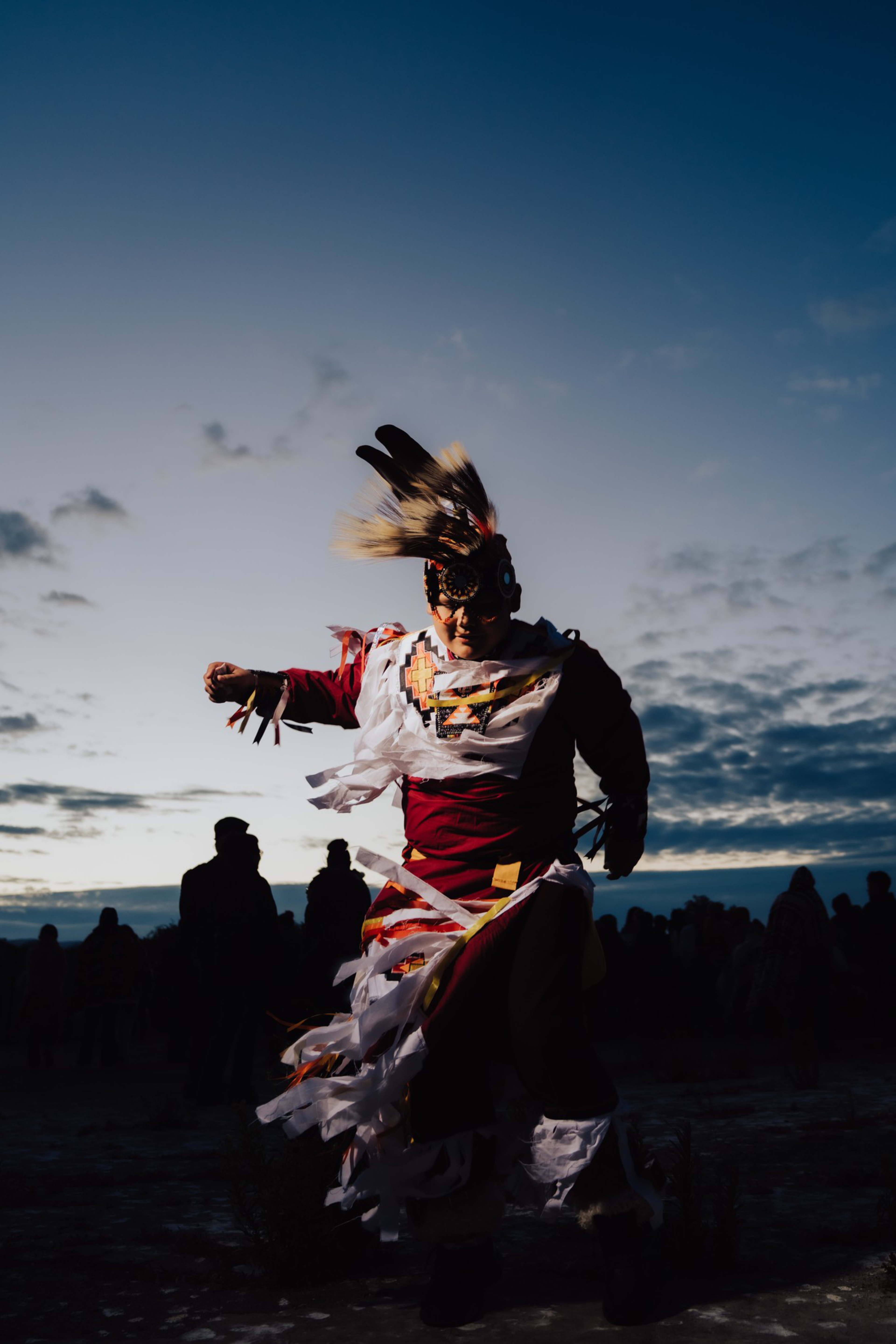 A dancer dressed in vibrant traditional attire with feathers and ribbons moves energetically against a dusky sky, surrounded by silhouetted onlookers.