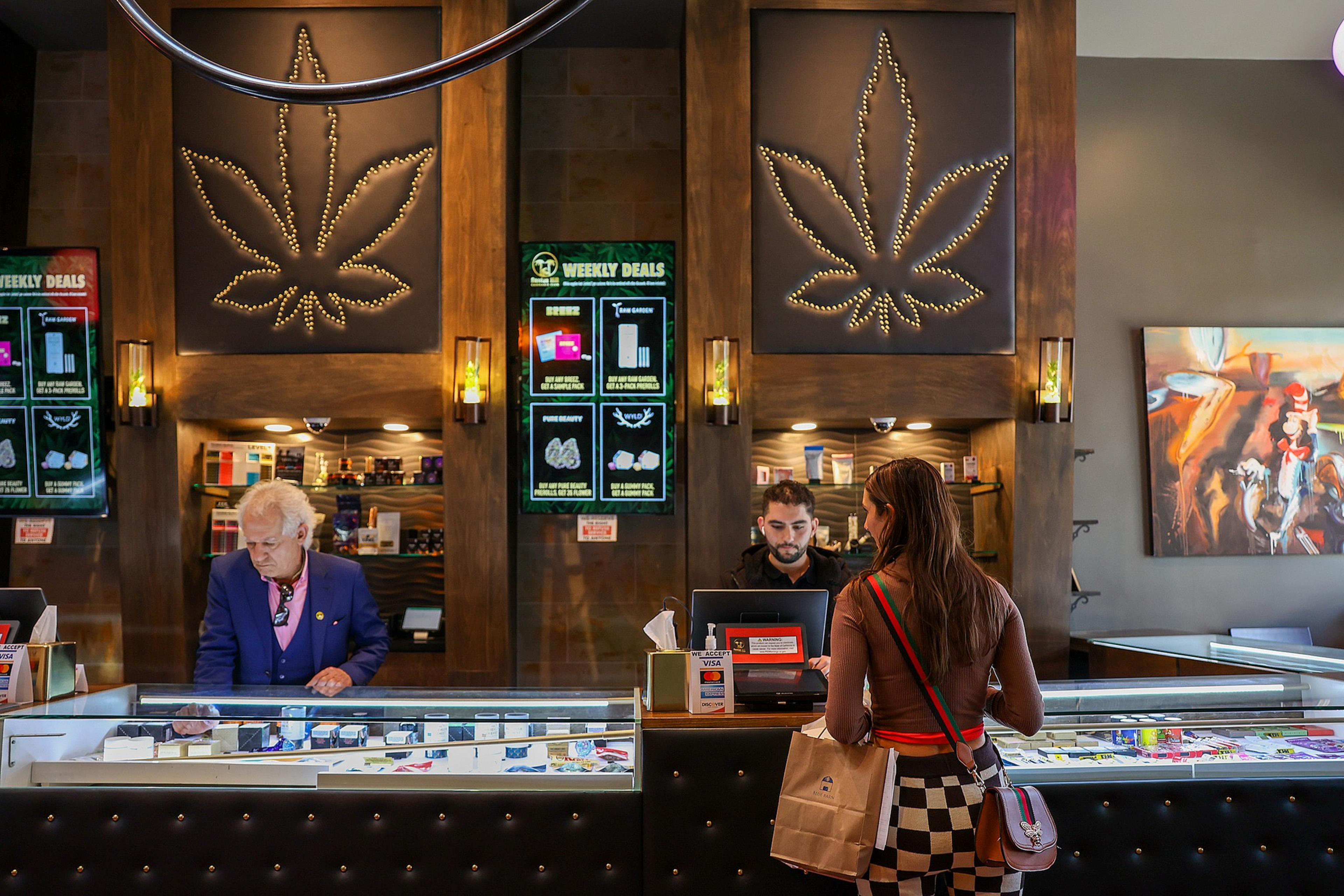 A dispensary counter with two employees, a customer, digital screens listing deals, and large illuminated cannabis leaf decorations on the wall.