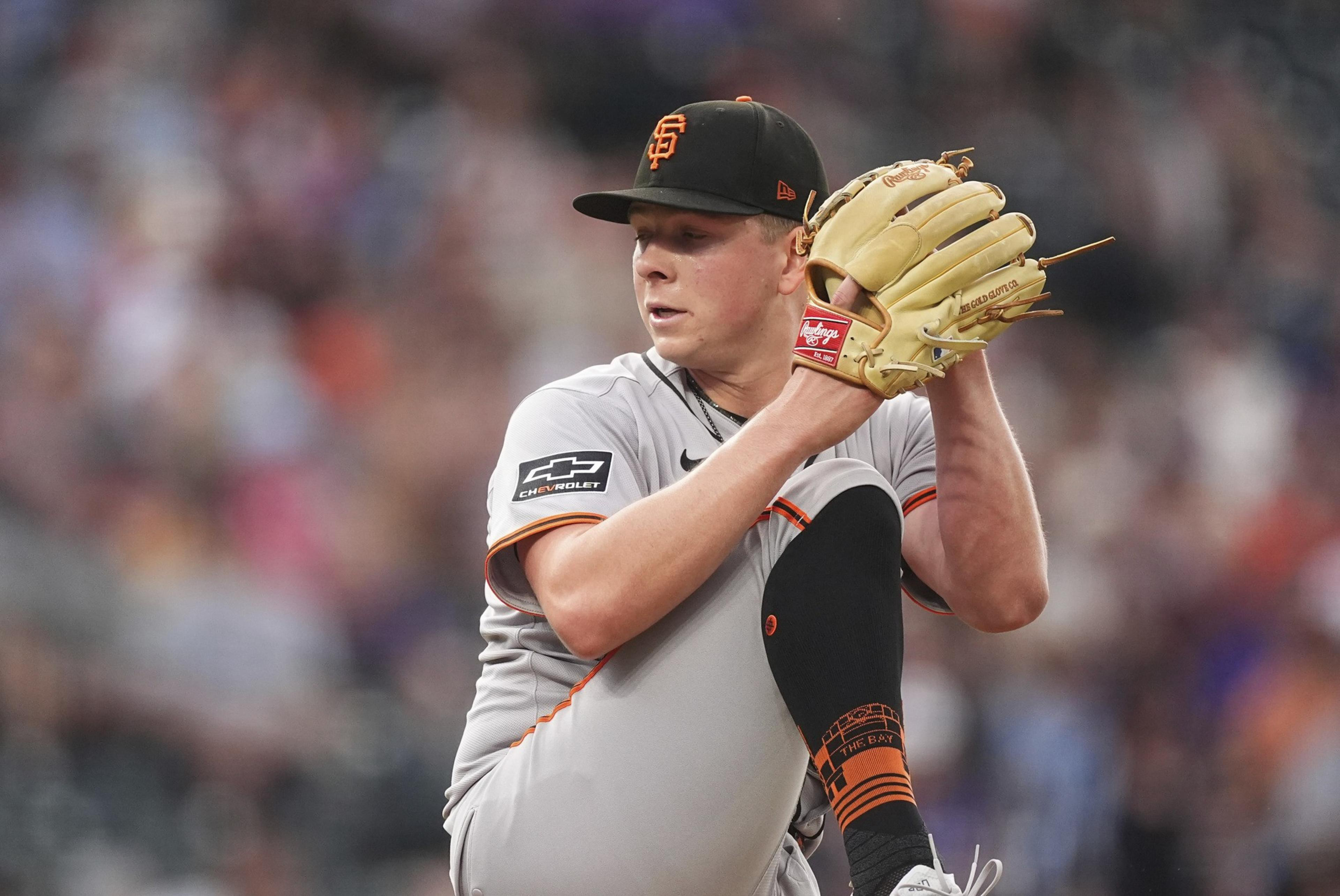A baseball player in a gray uniform and black cap is pitching, holding a glove, with one knee raised. He's focused, with a blurred crowd behind him.
