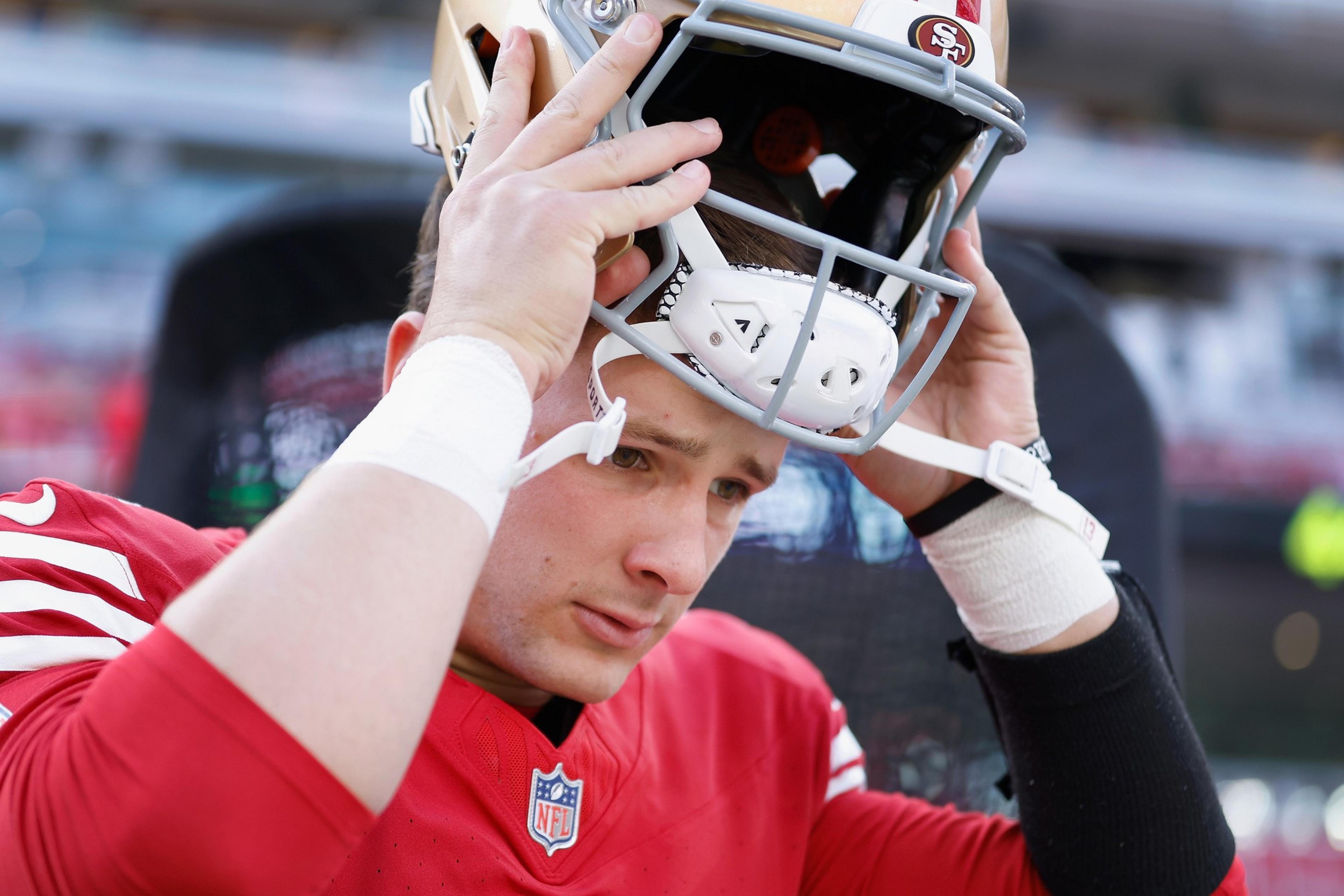 A football player in a red jersey is putting on a helmet. His expression is focused, and he's wearing white wristbands. The background is blurred.