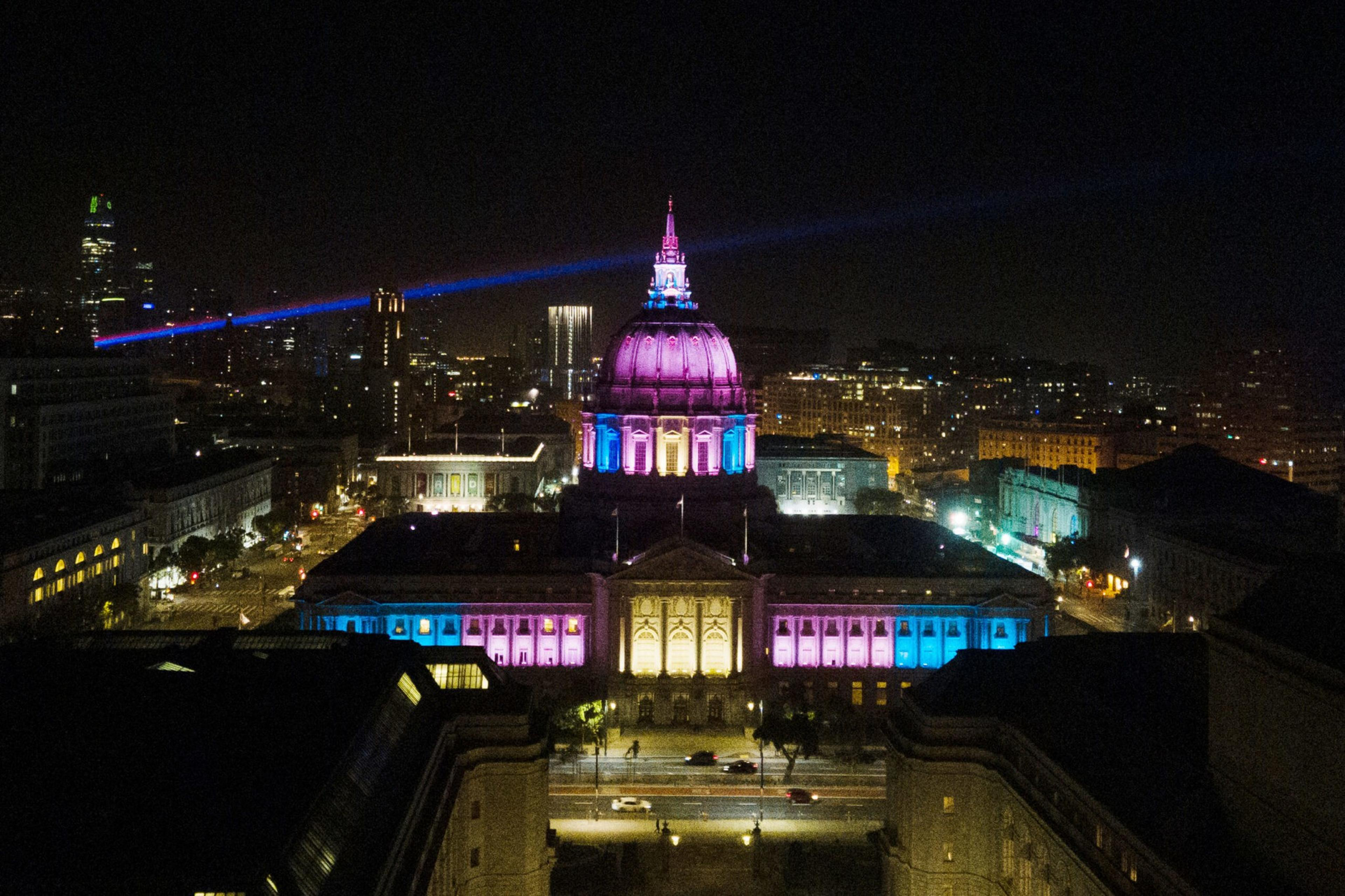 A large dome-shaped building is illuminated with purple, blue, and pink lights at night, with city lights and buildings visible in the background.