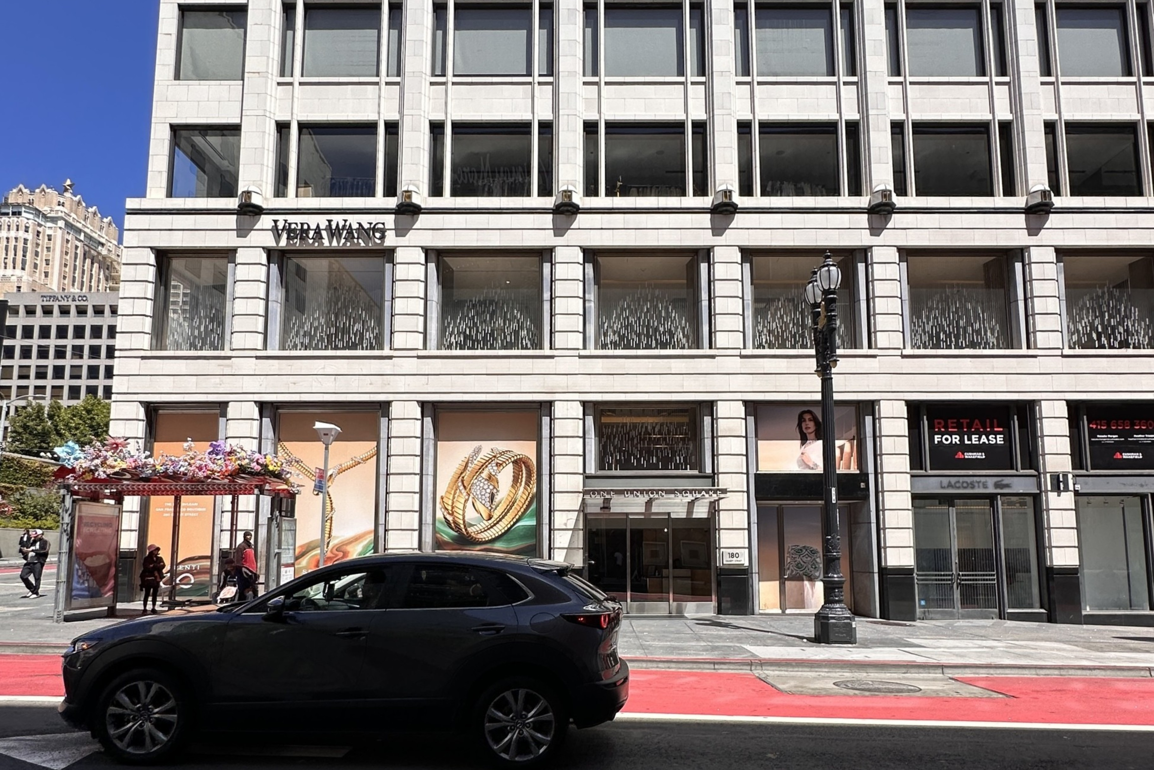 A multi-story white building with large windows features a Vera Wang store, a bus stop with floral decorations, a black SUV, and a red bike lane in front.