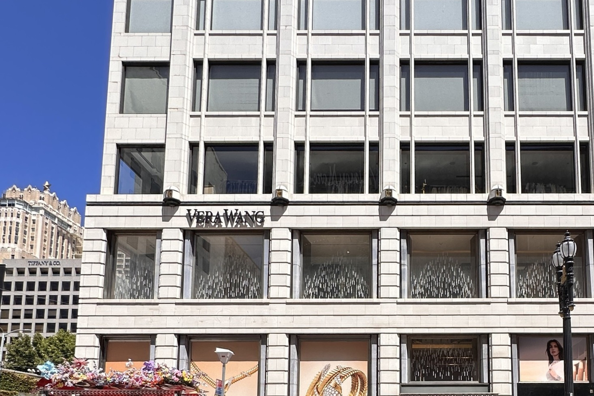 A multi-story white building with large windows features a Vera Wang store, a bus stop with floral decorations, a black SUV, and a red bike lane in front.
