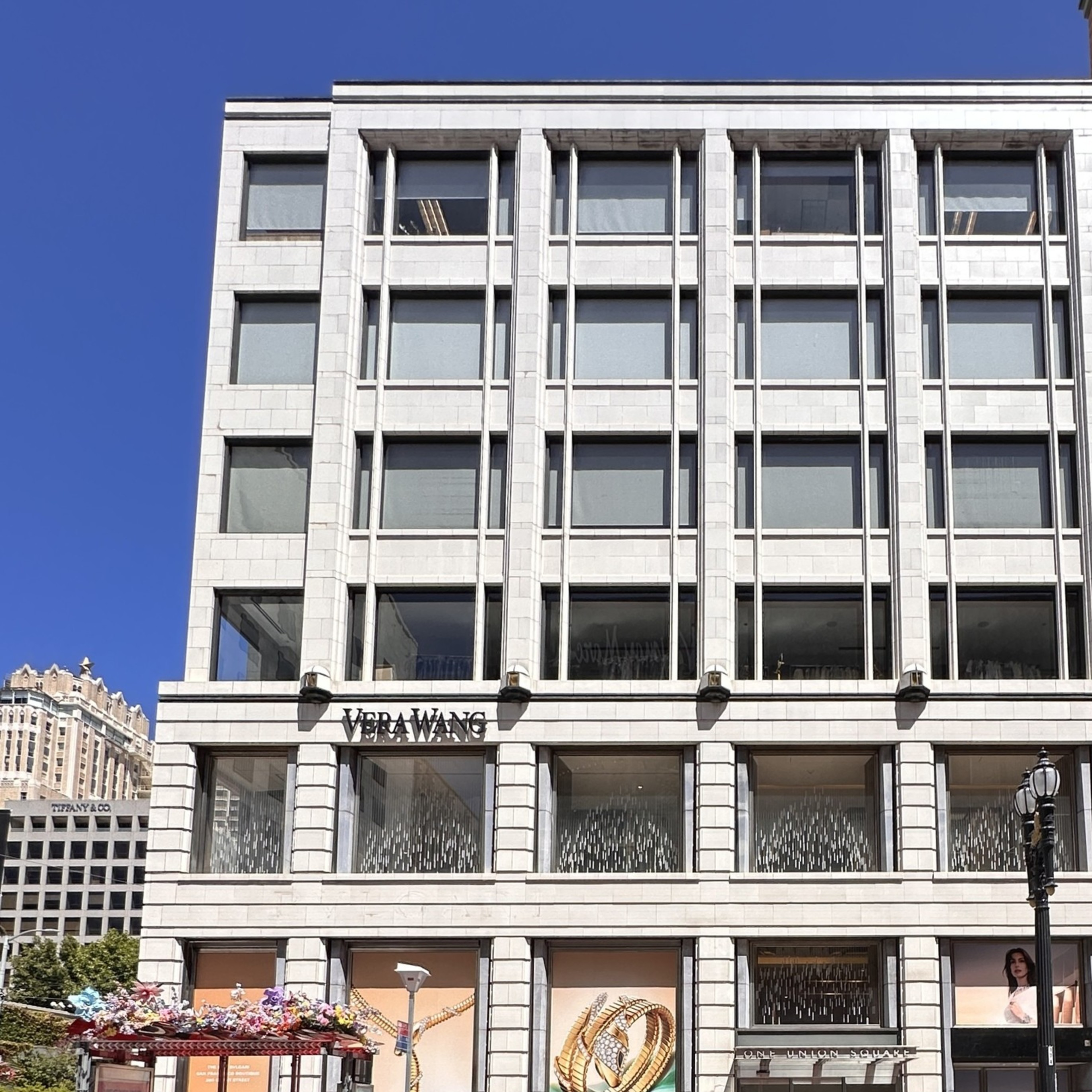 A multi-story white building with large windows features a Vera Wang store, a bus stop with floral decorations, a black SUV, and a red bike lane in front.