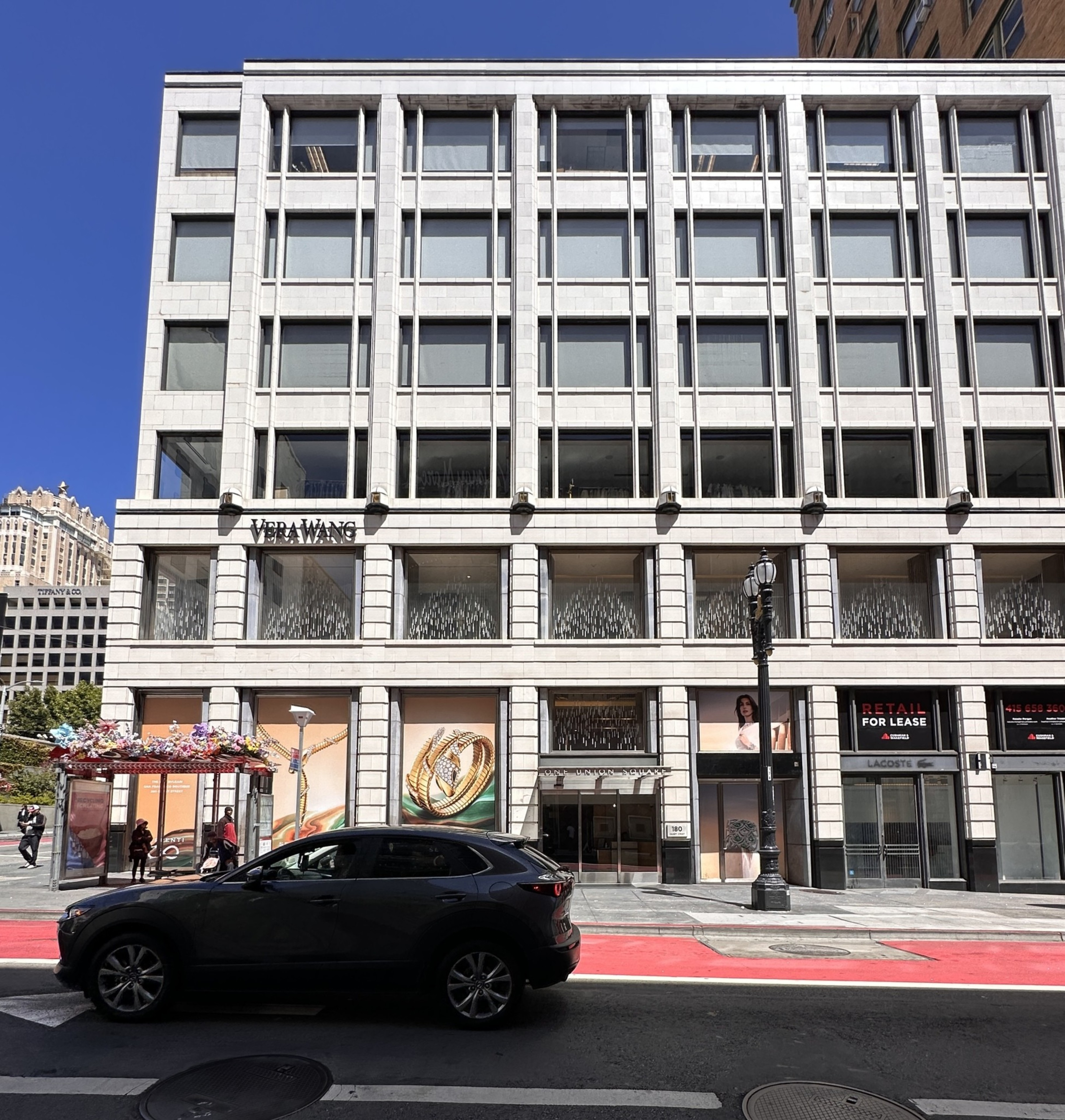 A multi-story white building with large windows features a Vera Wang store, a bus stop with floral decorations, a black SUV, and a red bike lane in front.
