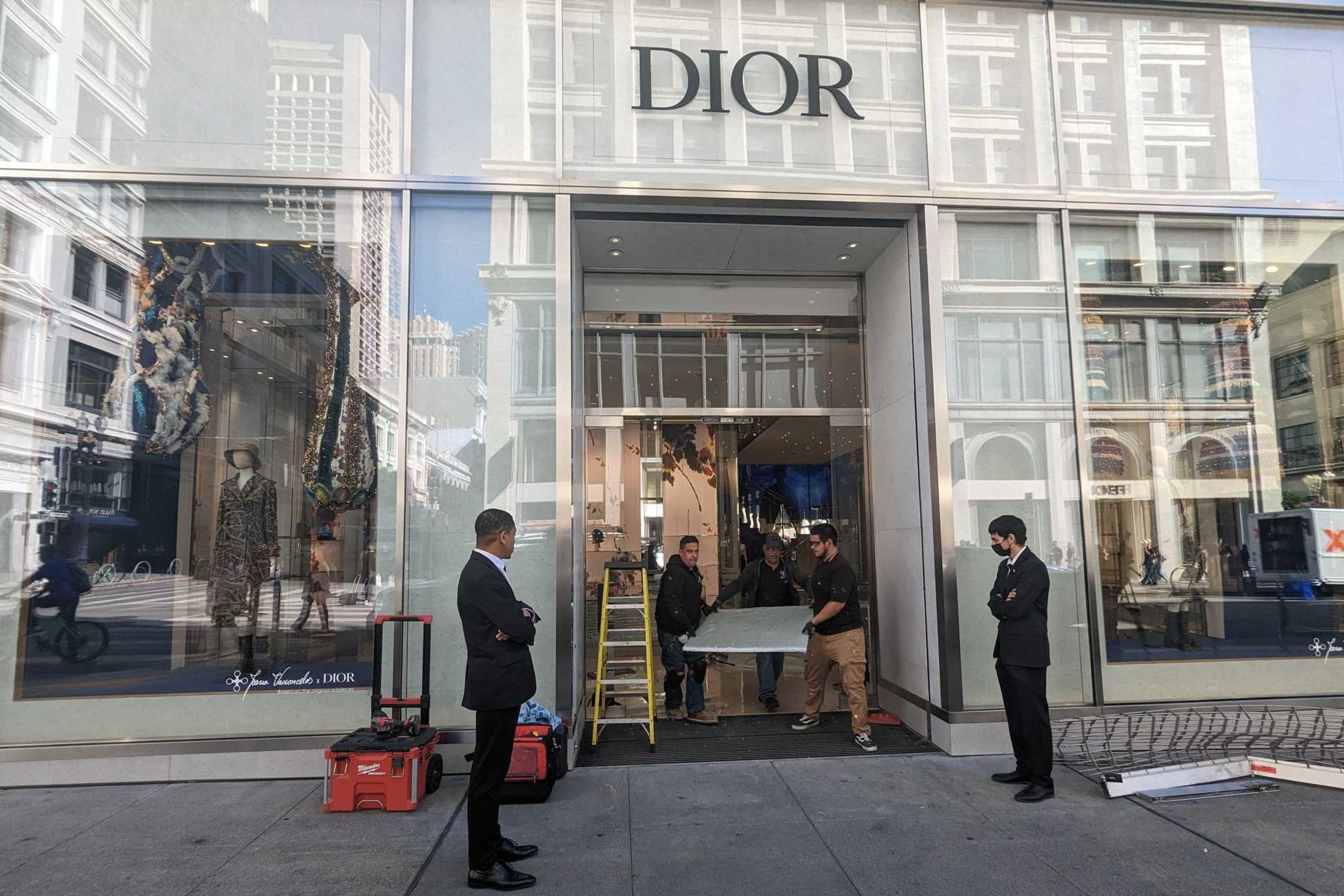 Security guards stand near the entrance to a luxury clothing store while workmen fix the doorway.