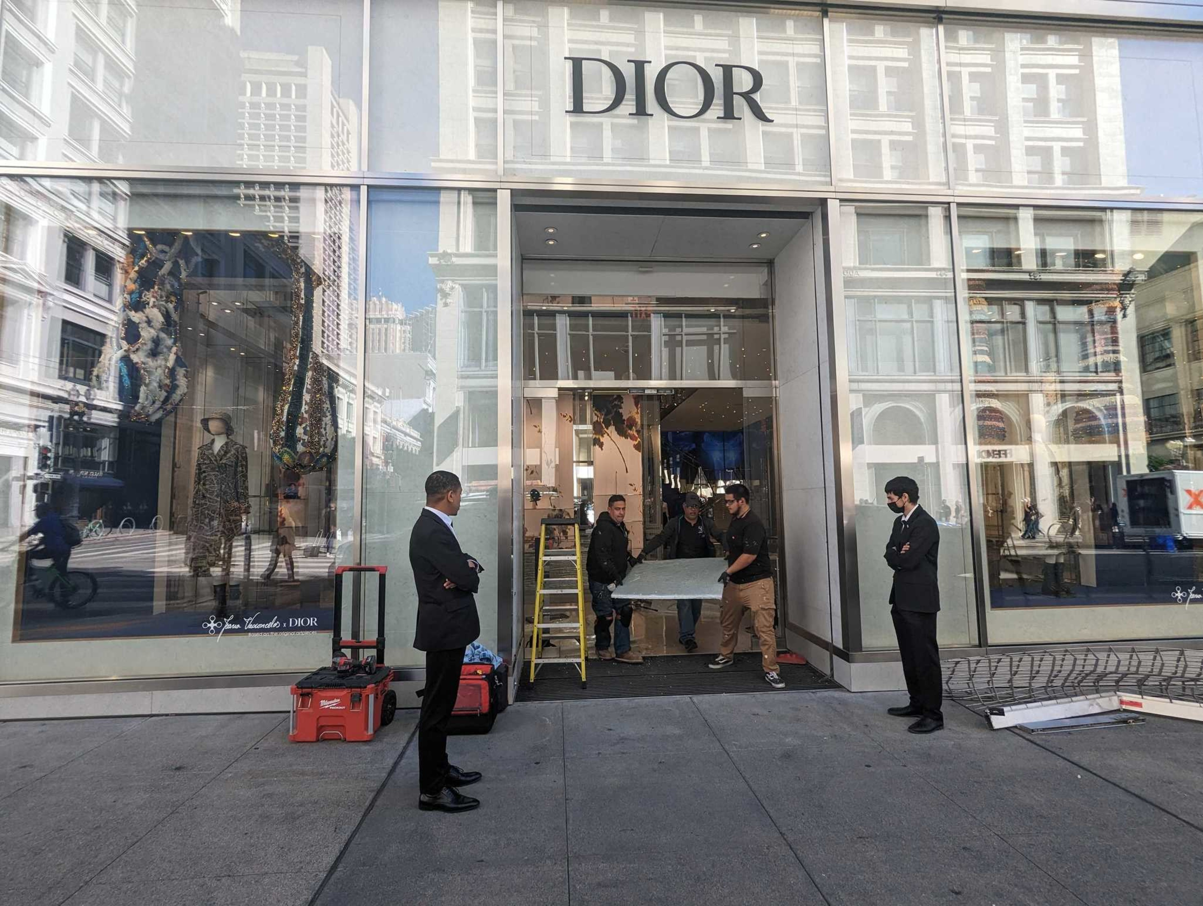 Security guards stand near the entrance to a luxury clothing store while workmen fix the doorway.