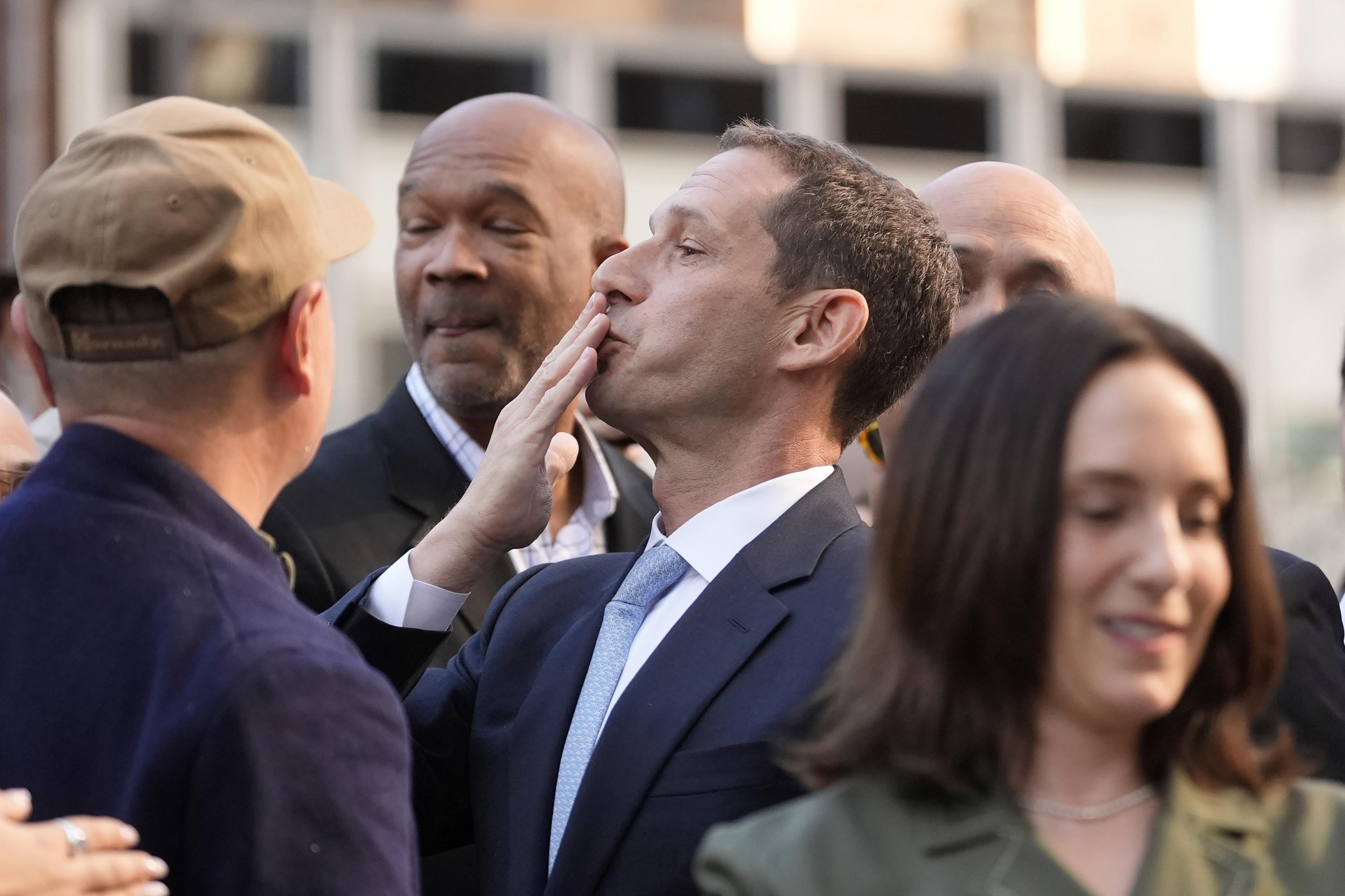 A man in a suit blows a kiss, surrounded by others in a crowded outdoor setting. He faces a person in a hat, with several people partially visible around them.