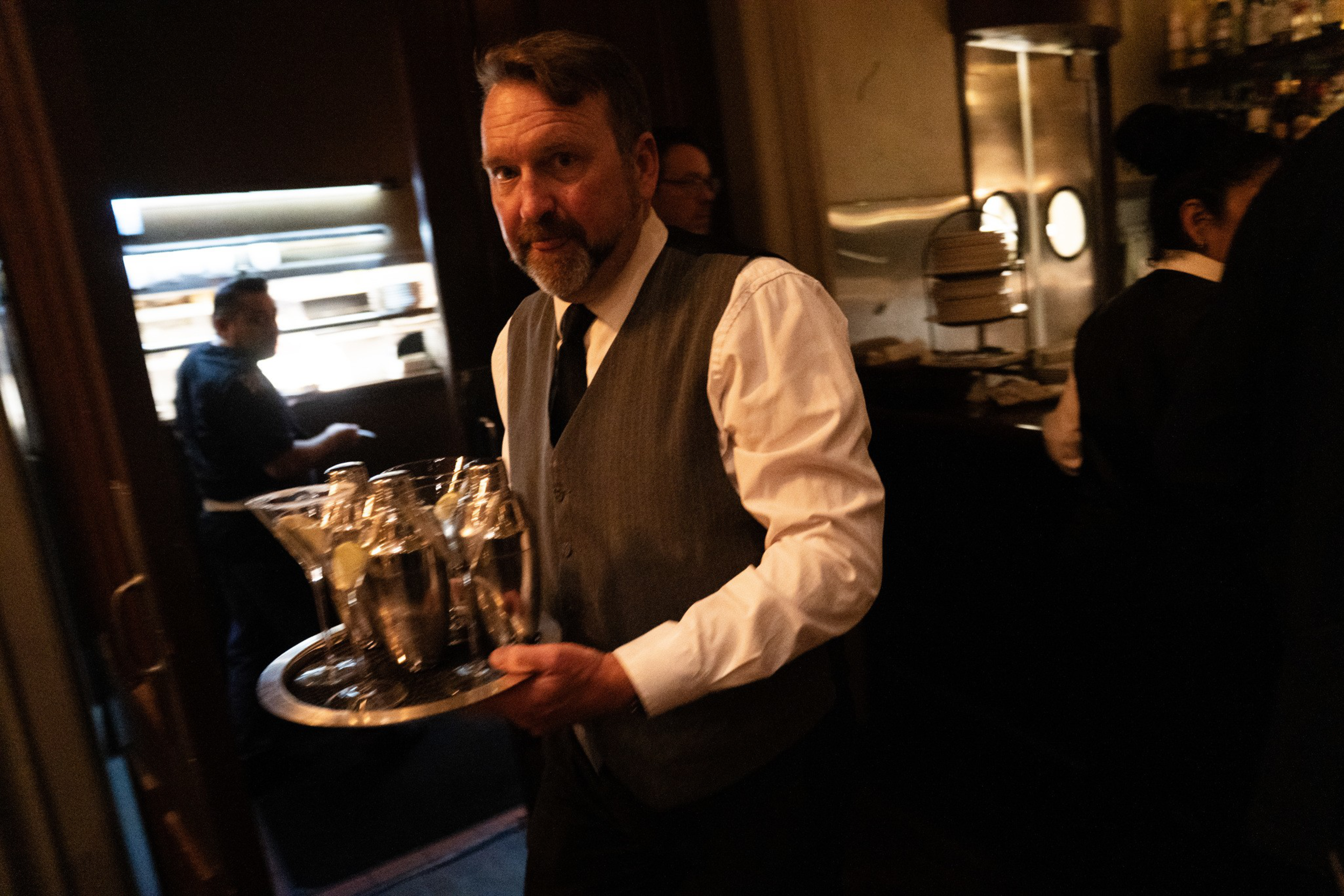 A man wearing a white shirt, vest, and tie carries a tray with cocktail shakers and glasses in a dimly lit bar or restaurant.
