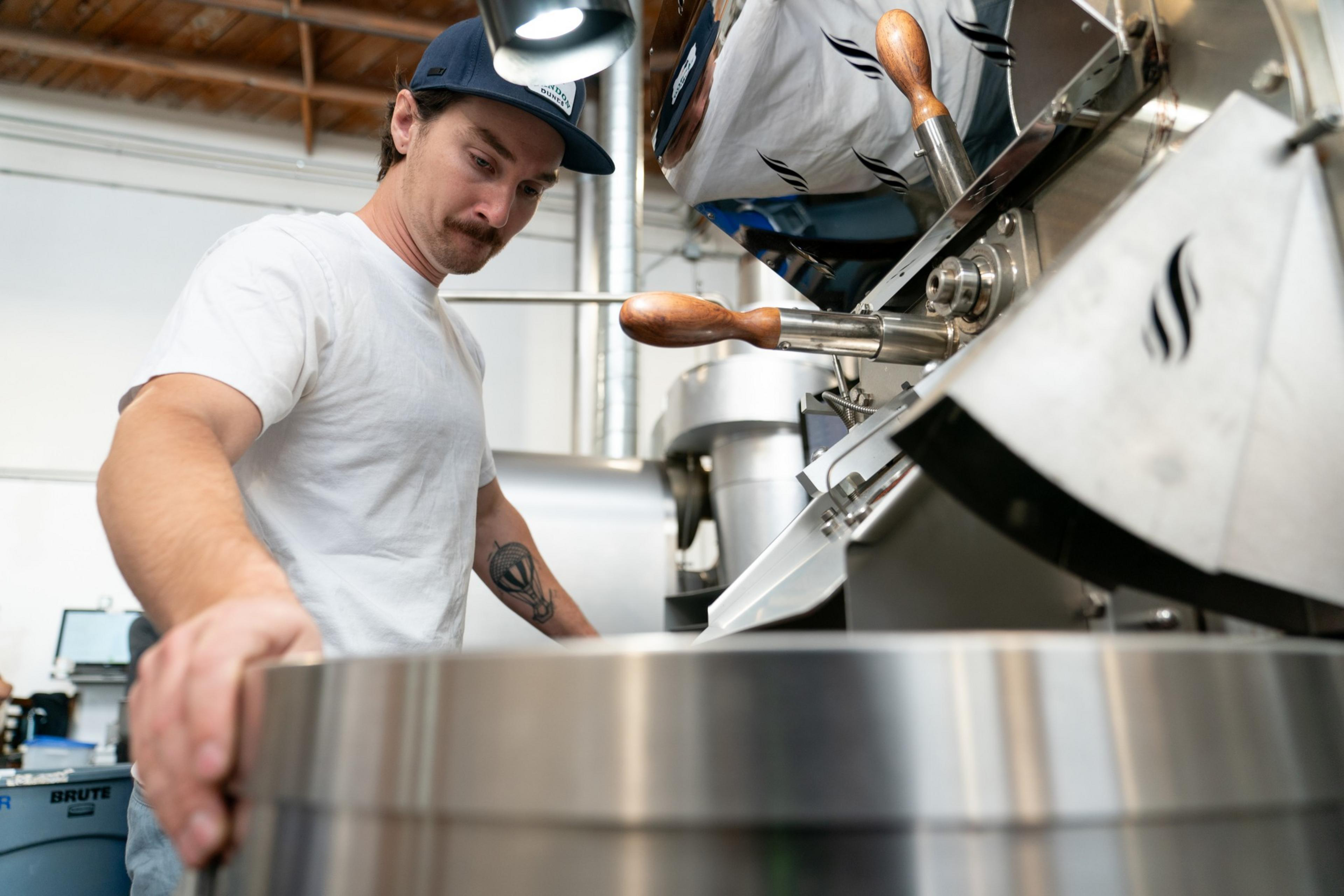 A man in a white shirt and navy cap operates a large, shiny coffee roasting machine in an industrial setting. His expression is focused as he adjusts the equipment.