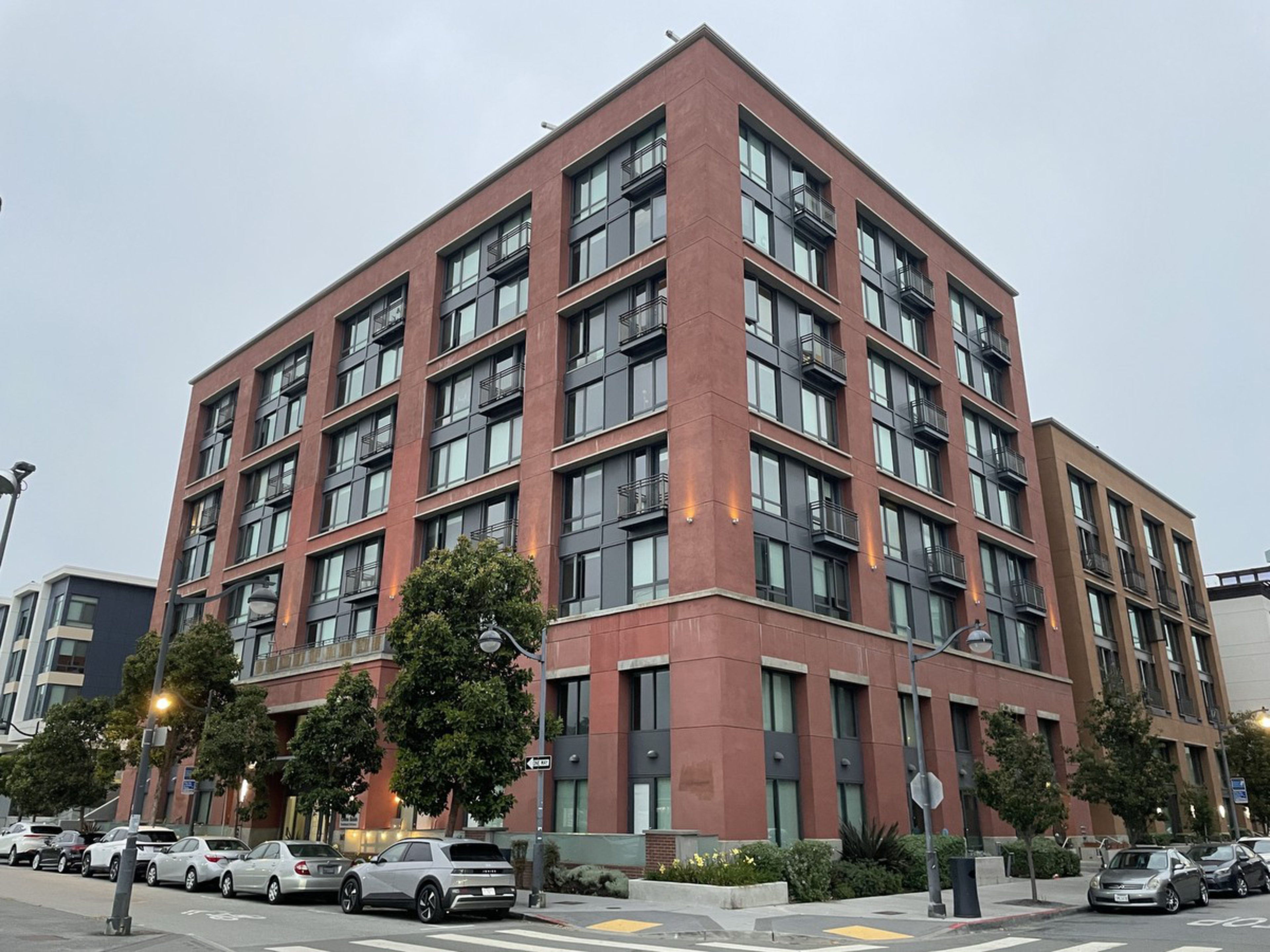 A modern five-story red brick and glass apartment building stands at a street corner with parked cars and trees lining the sidewalks.