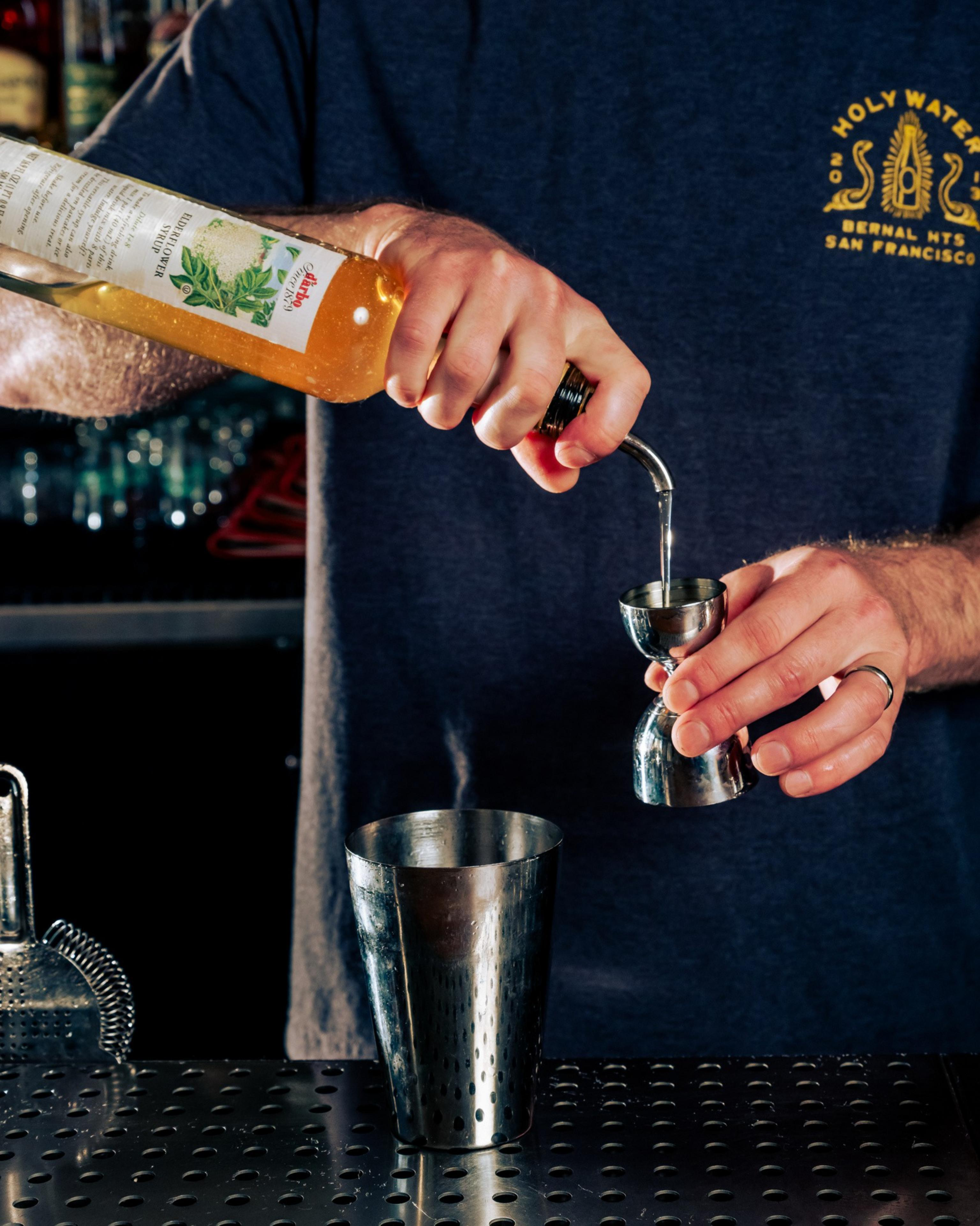 A person pours liquid from a bottle into a jigger above a cocktail shaker on a bar counter. They're wearing a dark T-shirt with a logo on it.