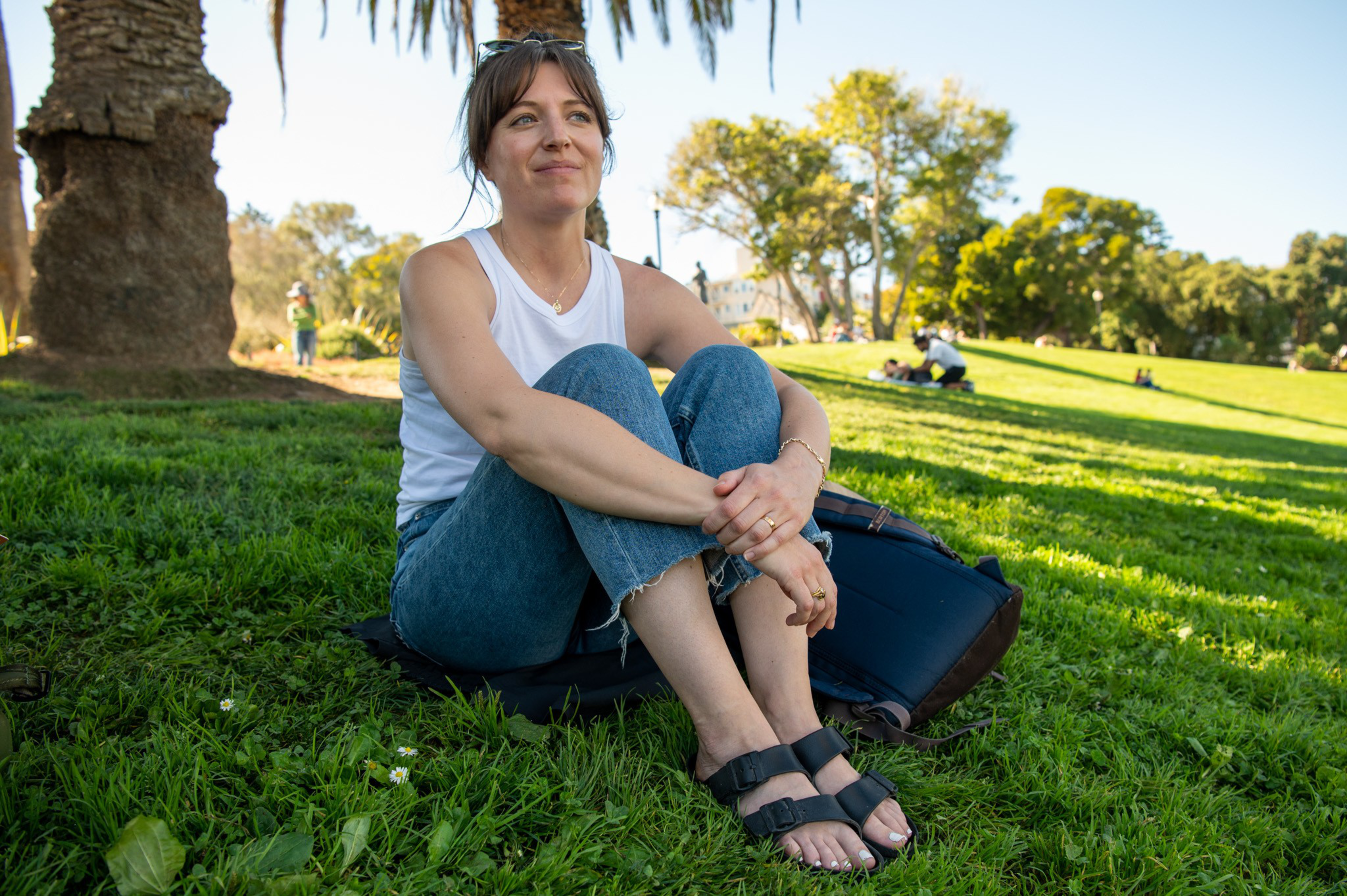 A woman with sunglasses on her head sits cross-legged on grass in a park, wearing a white tank top, blue jeans, and black sandals.