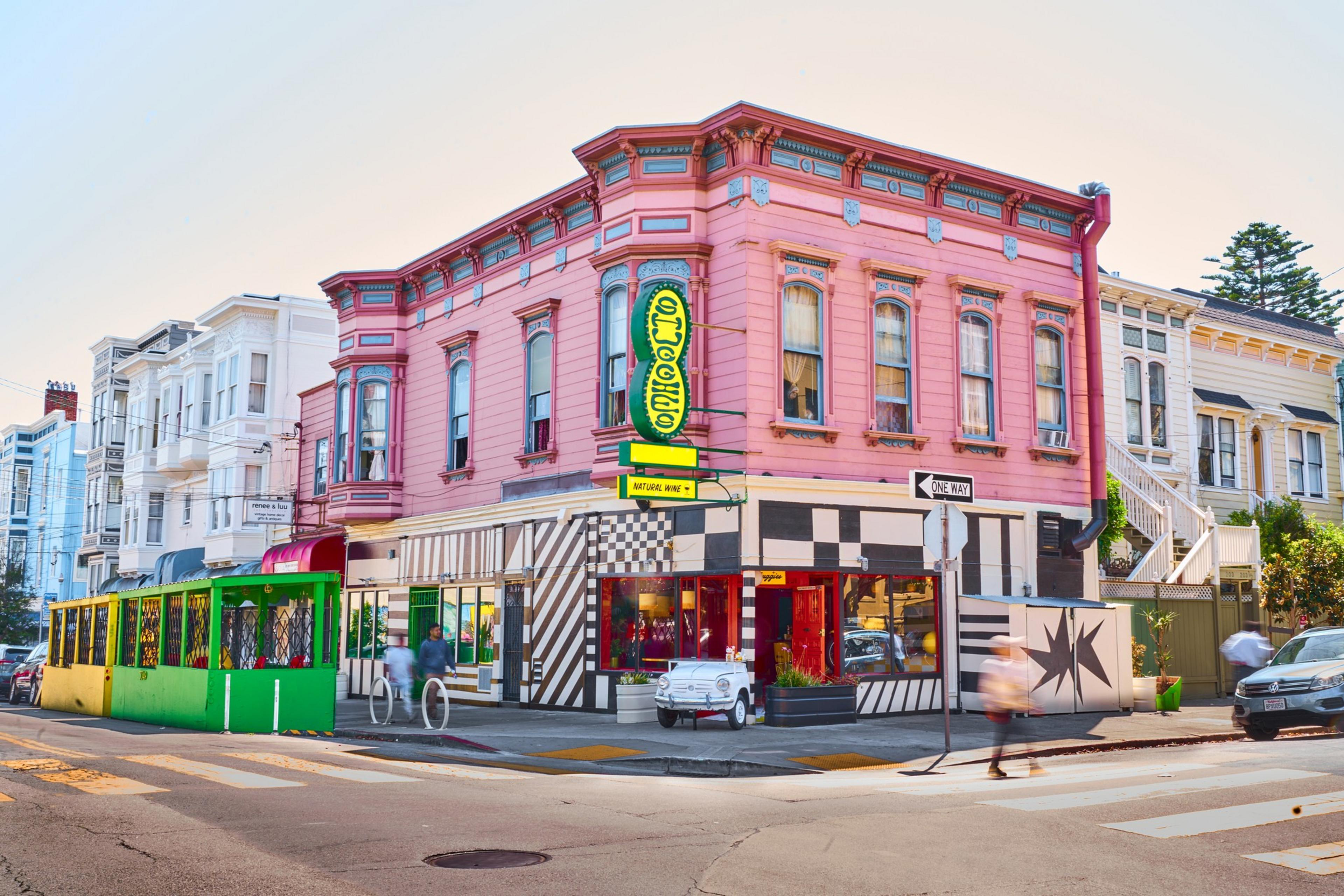 A pink two-story corner building with a black-and-white striped base and a green and yellow “Michocito” sign, with outdoor seating in a green enclosure.