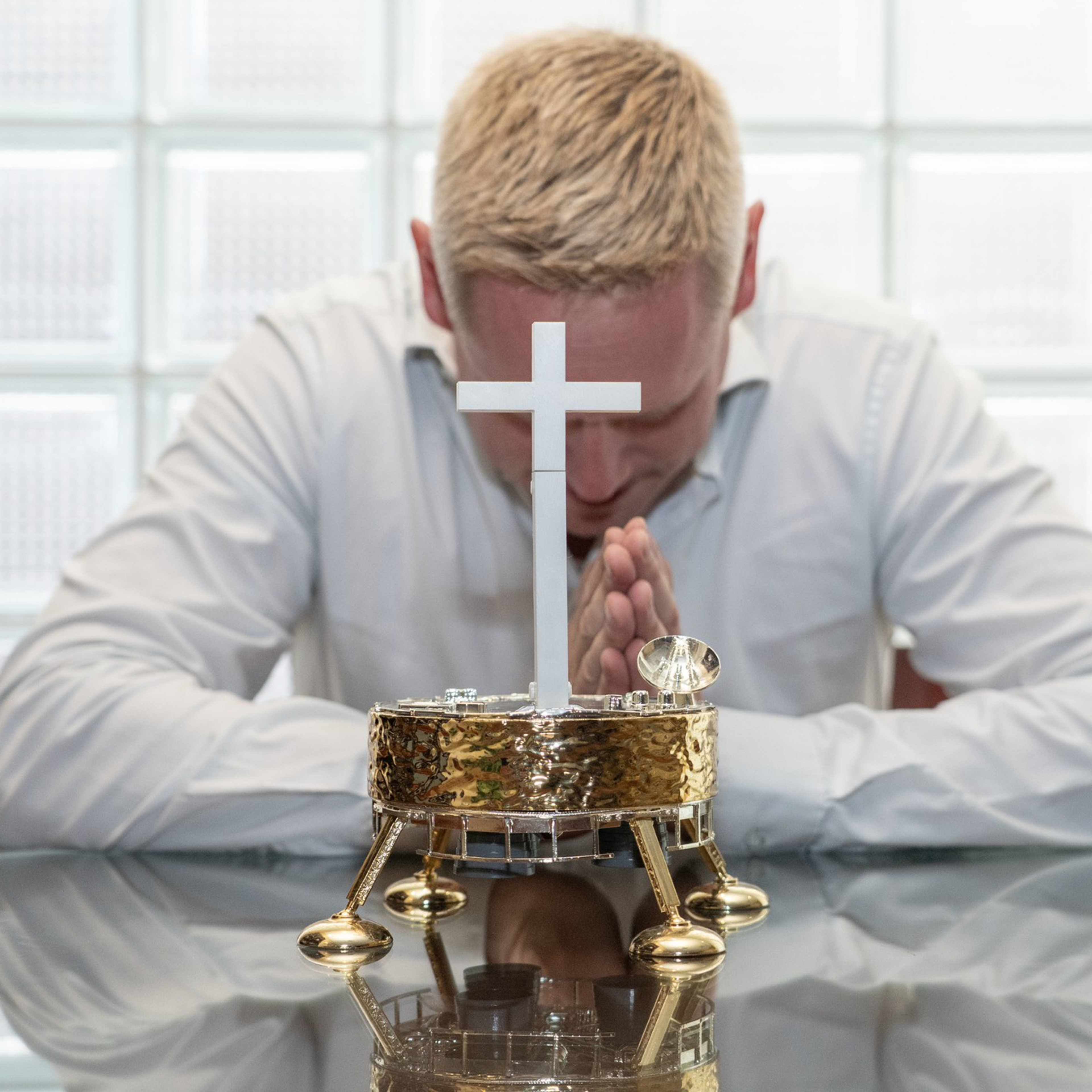 A man with blond hair in a white shirt sits at a glass table, bowing his head and praying behind a small gold tabernacle with a white cross.