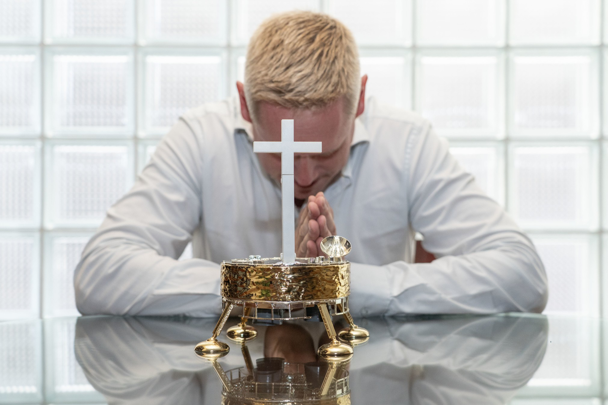 A man with blond hair in a white shirt sits at a glass table, bowing his head and praying behind a small gold tabernacle with a white cross.