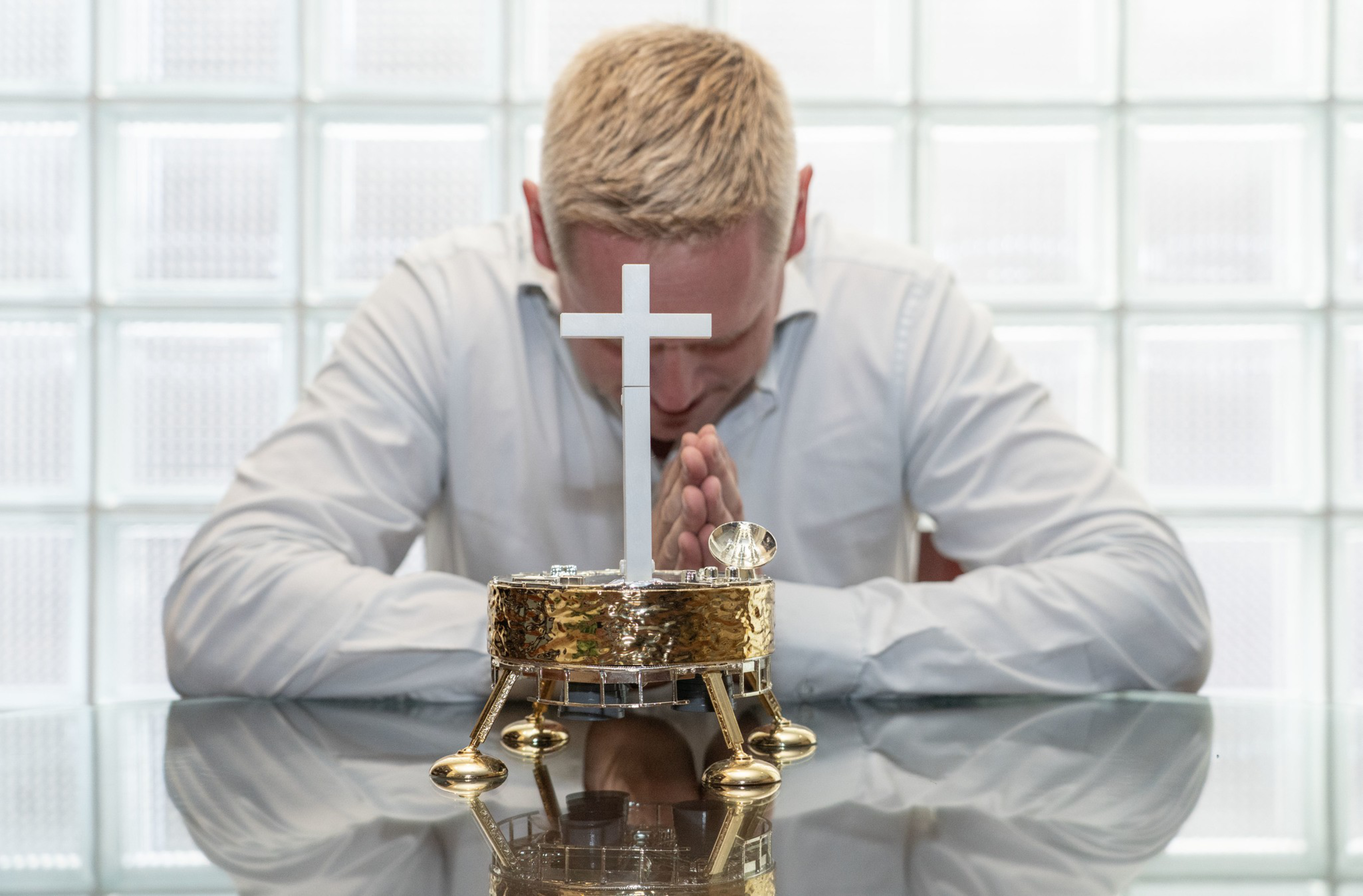A man with blond hair in a white shirt sits at a glass table, bowing his head and praying behind a small gold tabernacle with a white cross.