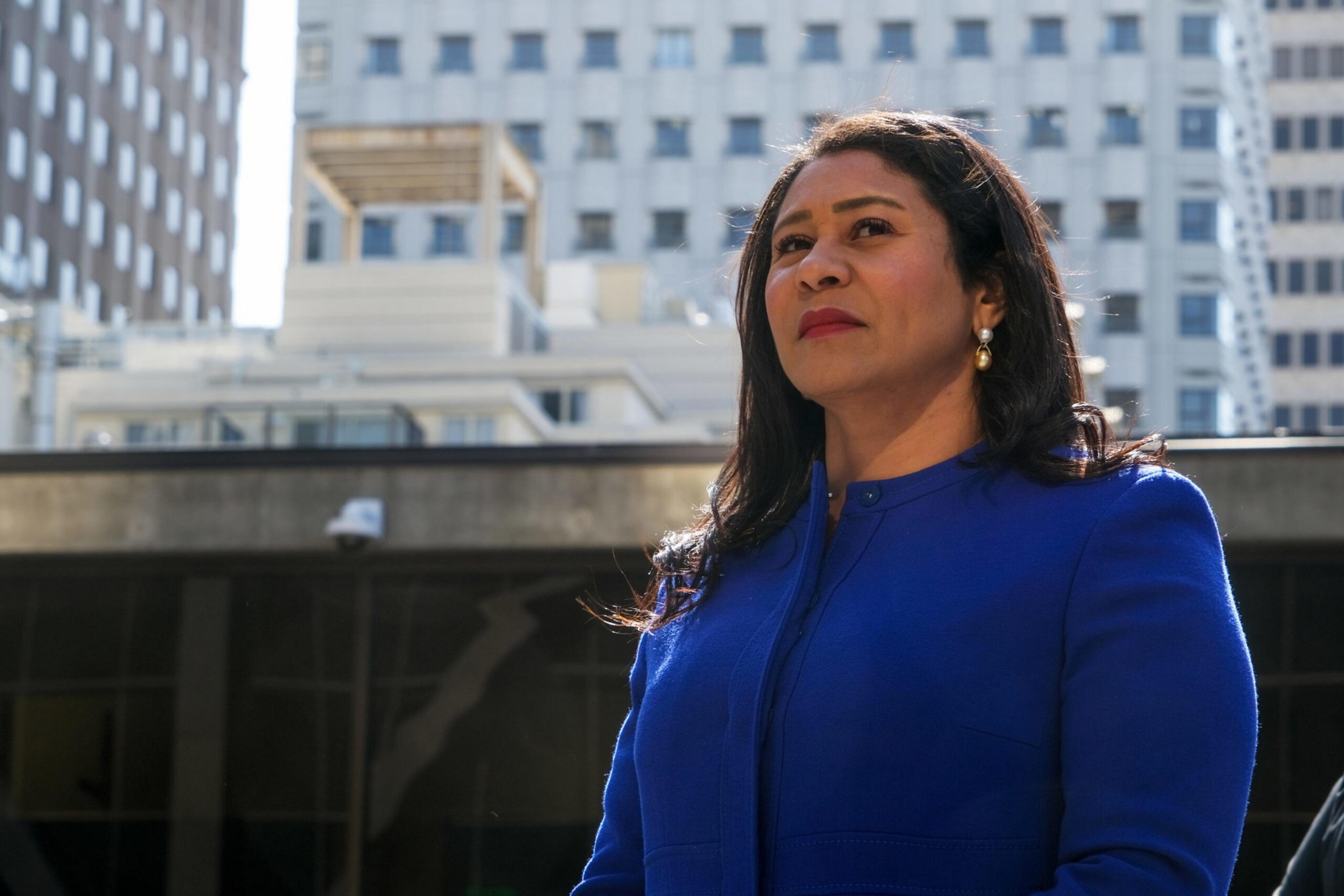 A woman in a royal blue coat and pearl earrings stands confidently outdoors with tall buildings in the background under bright daylight.