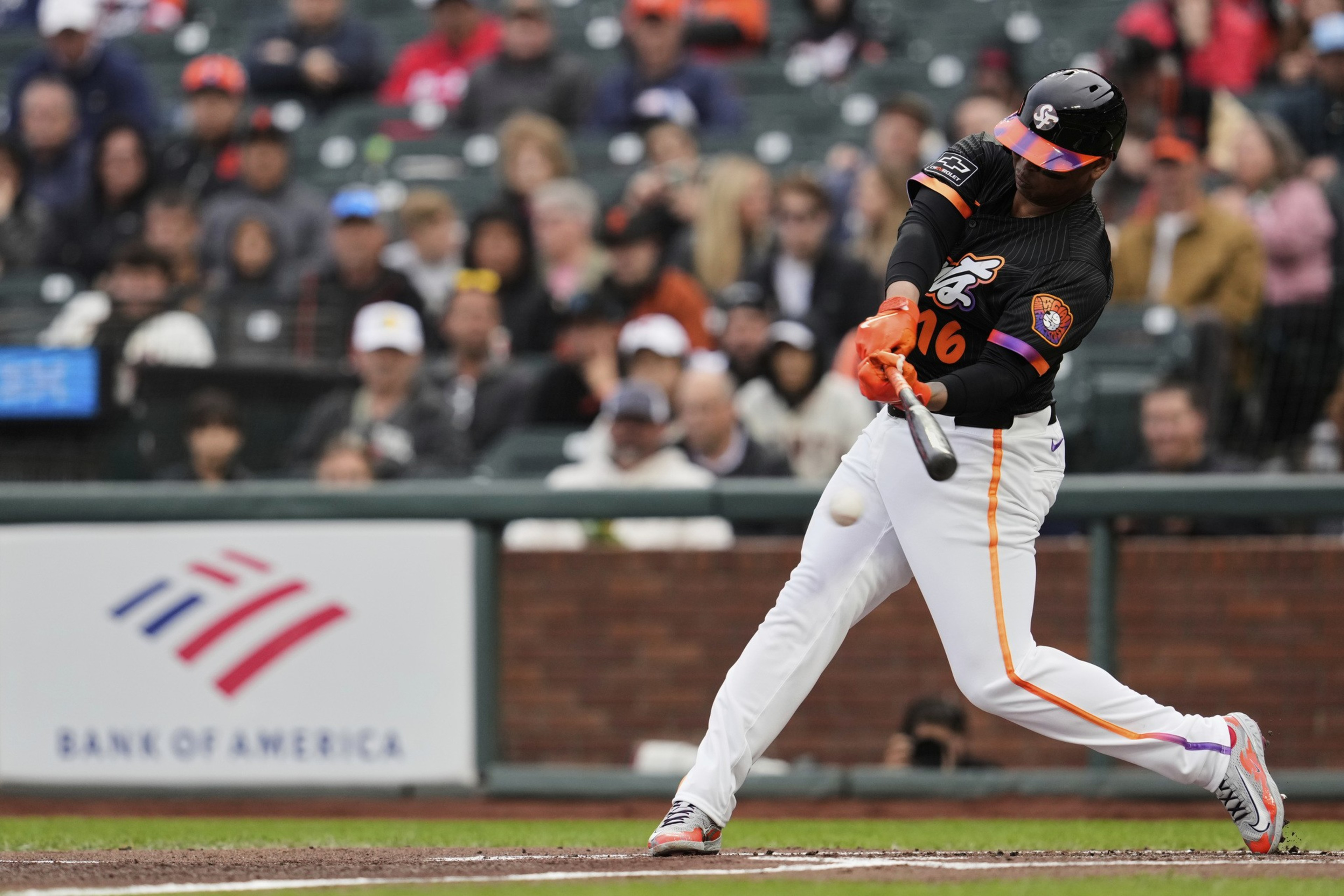 A baseball player in black and white uniform swings a bat, hitting the ball. Spectators in the background watch from the stands.