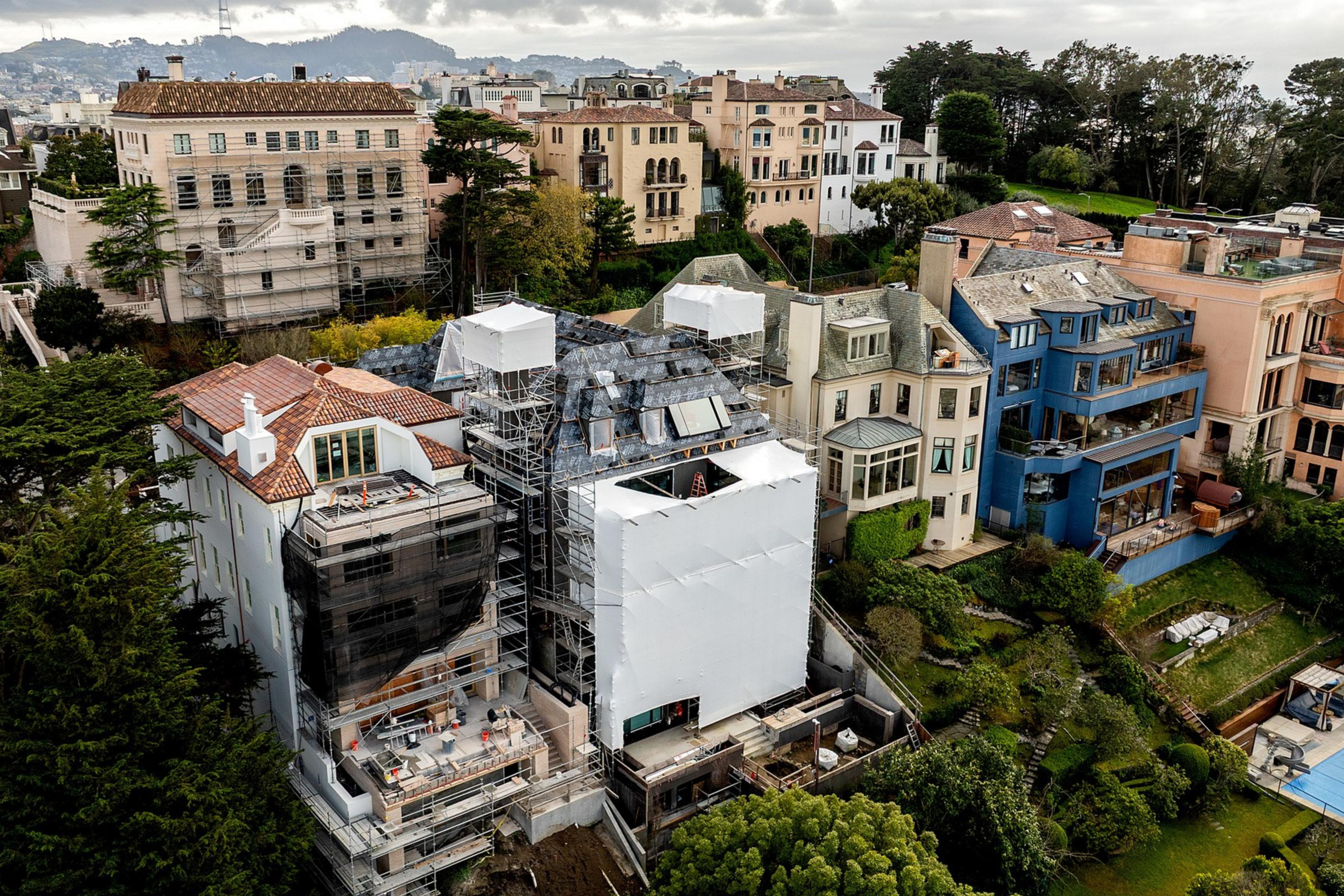 The image shows a cluster of large homes on a hillside, with one under renovation covered in scaffolding and white sheeting. Lush greenery surrounds the area.