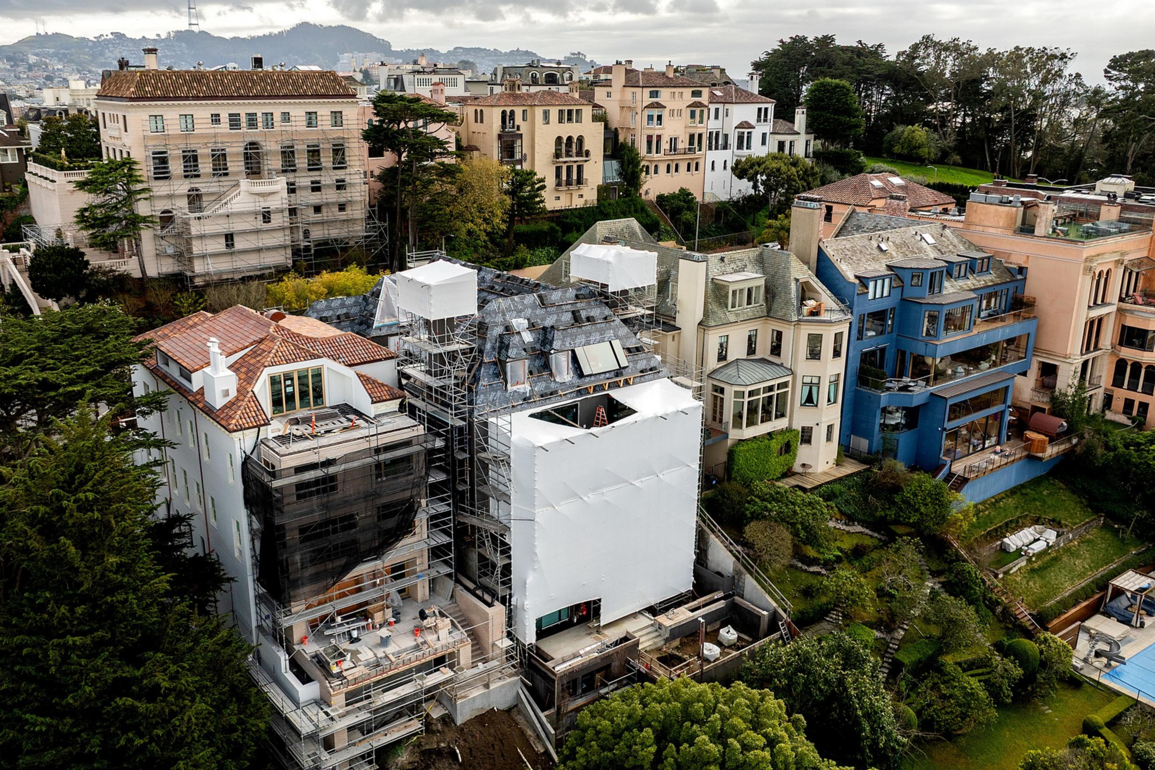 The image shows a cluster of large homes on a hillside, with one under renovation covered in scaffolding and white sheeting. Lush greenery surrounds the area.