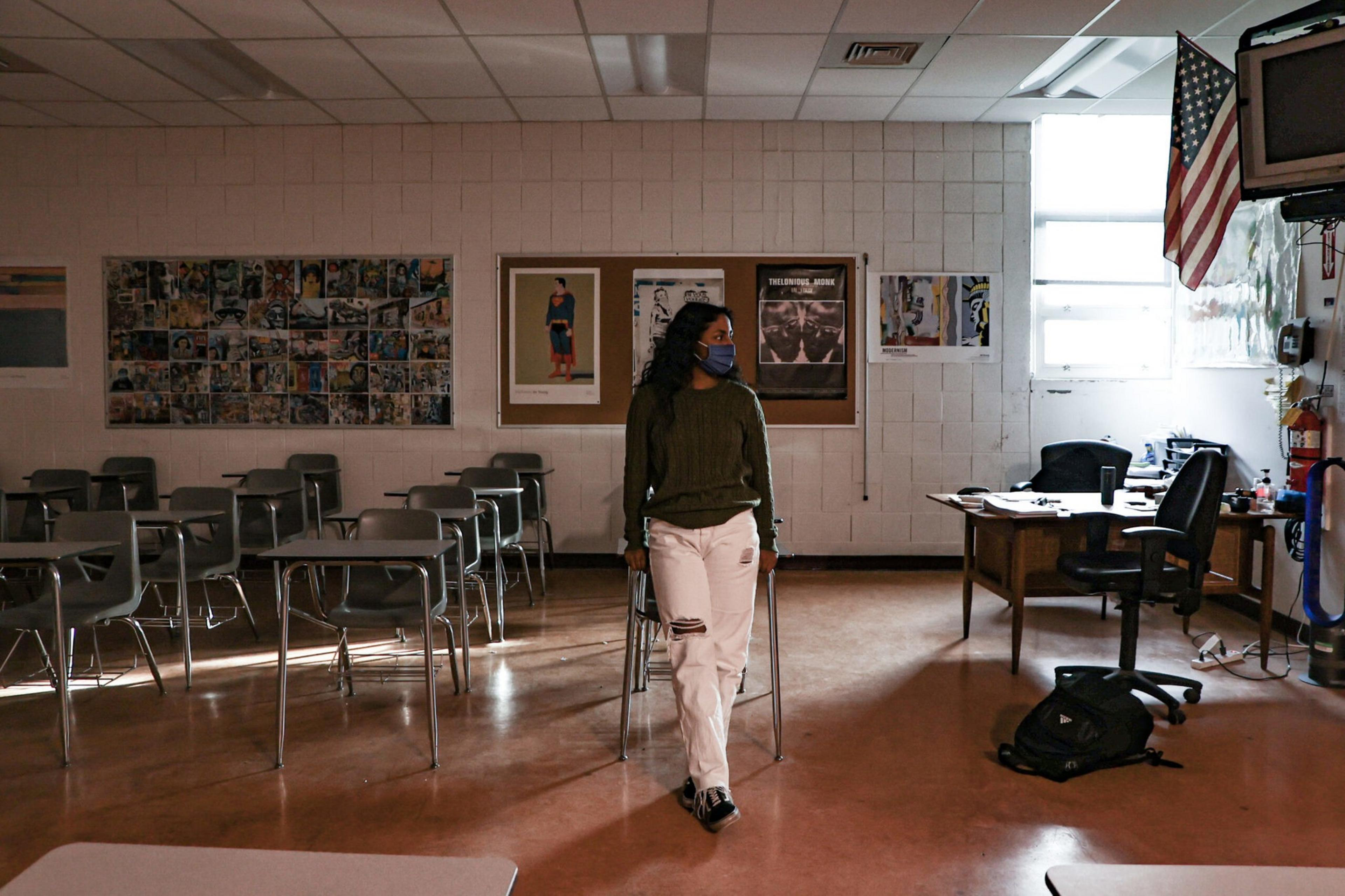 A person wearing a mask, green sweater, and ripped white pants stands in an empty classroom near desks, with an American flag and a teacher's desk in view.